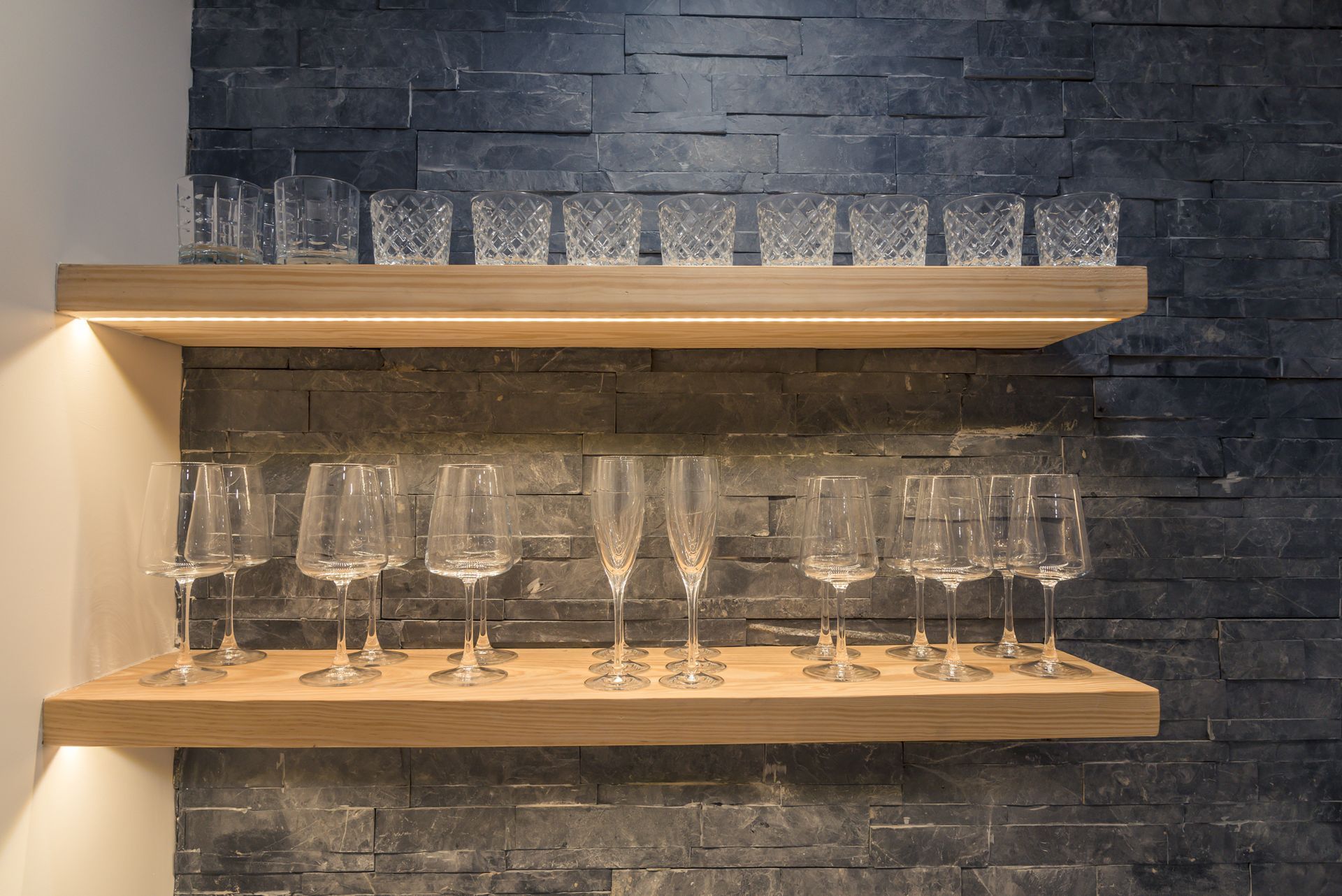Two wooden shelves against a dark stone wall, displaying rows of empty wine glasses.