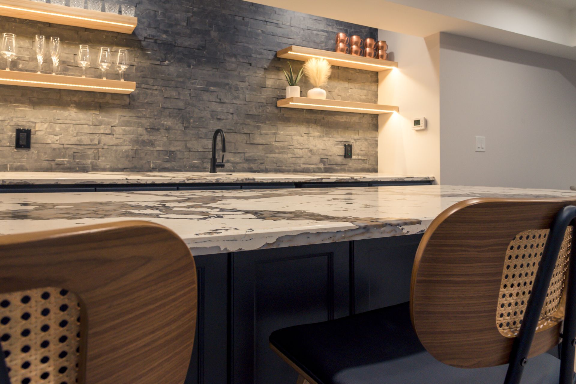Modern kitchen with marble island, dark cabinets, wood bar stools, and open shelves with glasses.