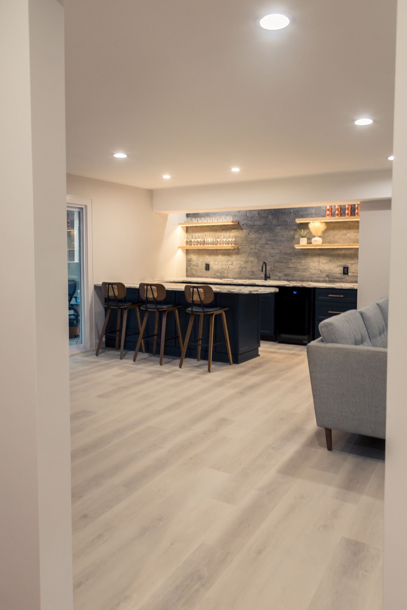 Modern open-plan living room with kitchen island, bar stools, stone backsplash, and gray sofa