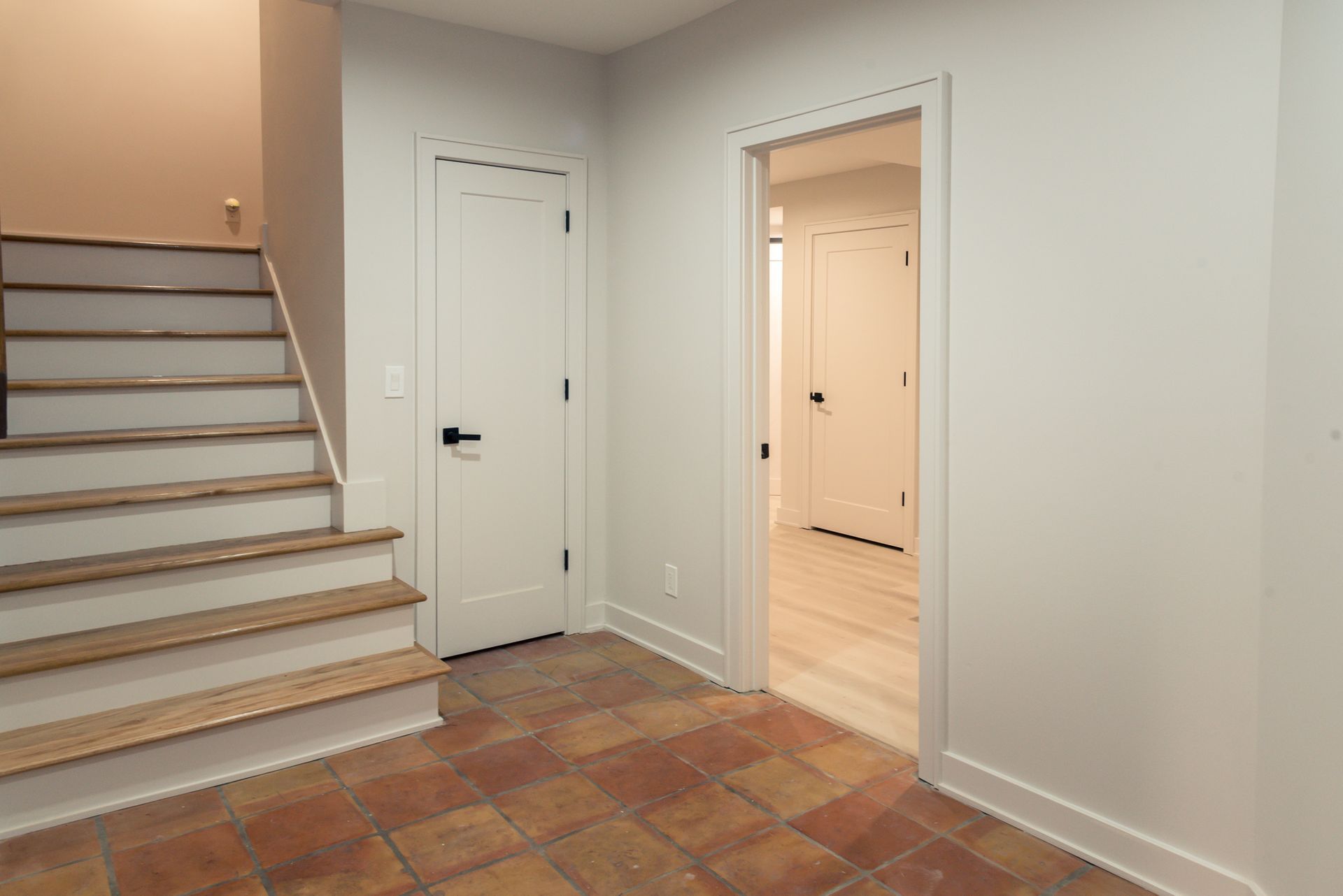 Interior hallway with stairs, white doors, tiled floor, and an open doorway to a lit room