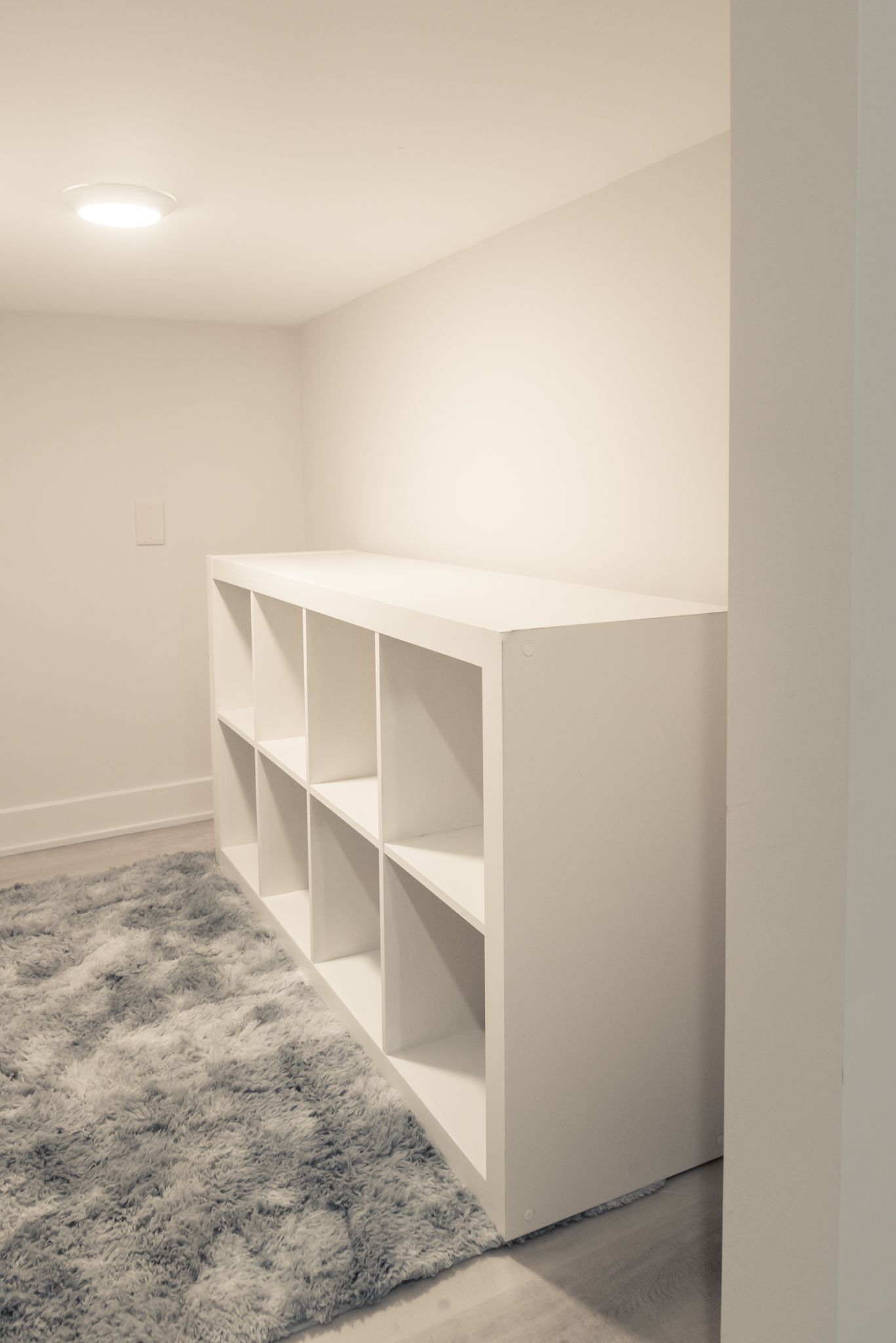 Minimal white shelving unit in a bright empty room with a gray shag rug and recessed ceiling light