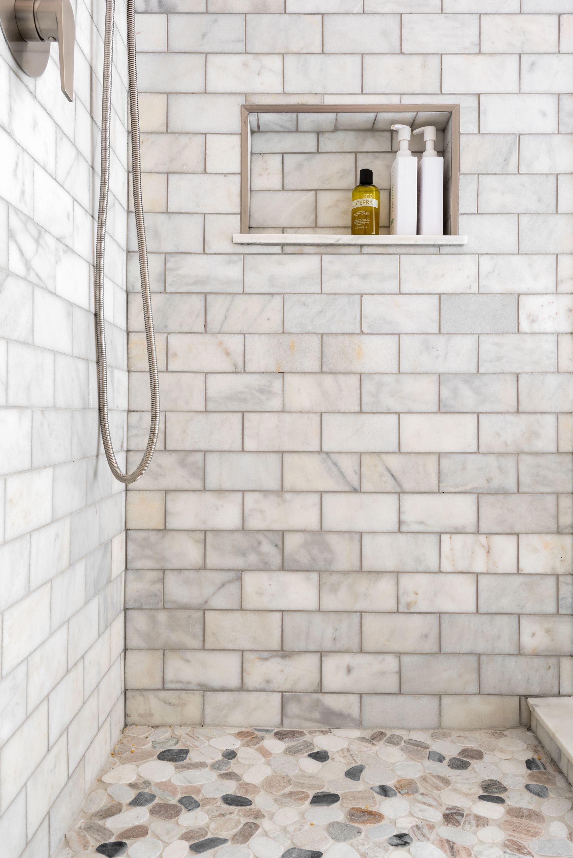 White tiled shower with a built-in shelf, hanging showerhead, and pebble stone floor.