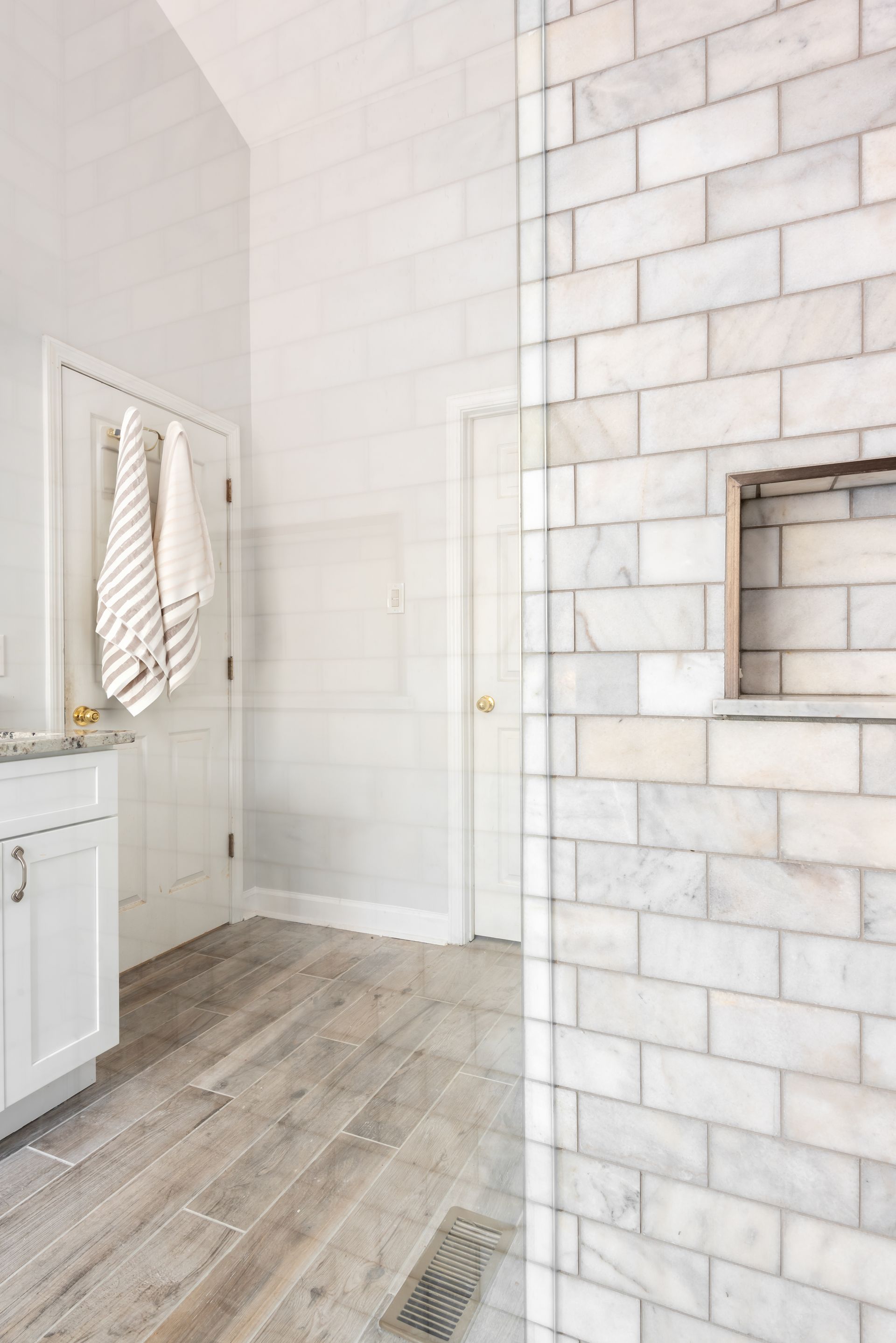 Bright bathroom with white tile, wood-look floor, vanity, towel, and a tiled shower wall with niche.