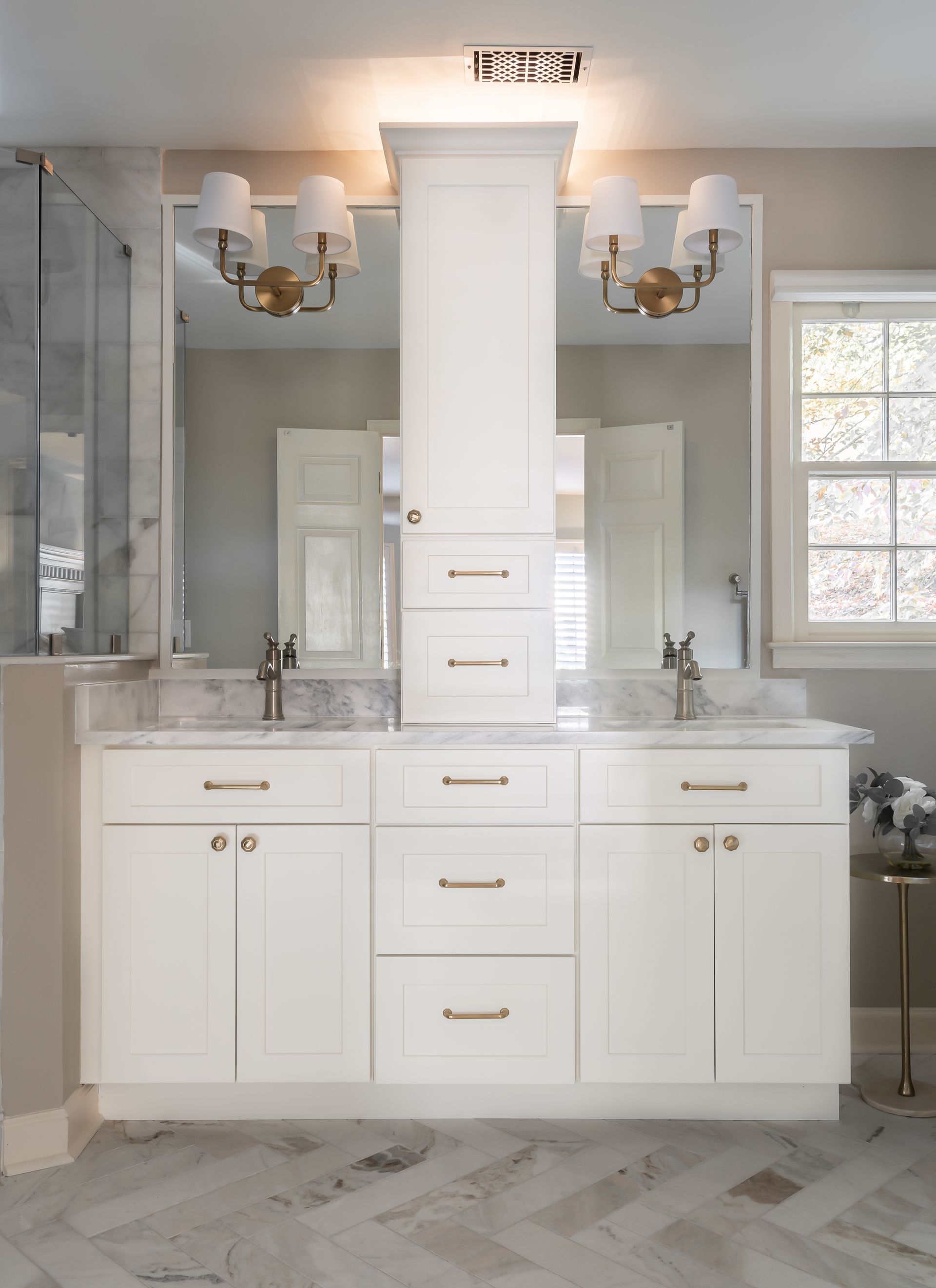 White bathroom vanity with dual sinks, large mirror, and brass sconces on marble flooring.