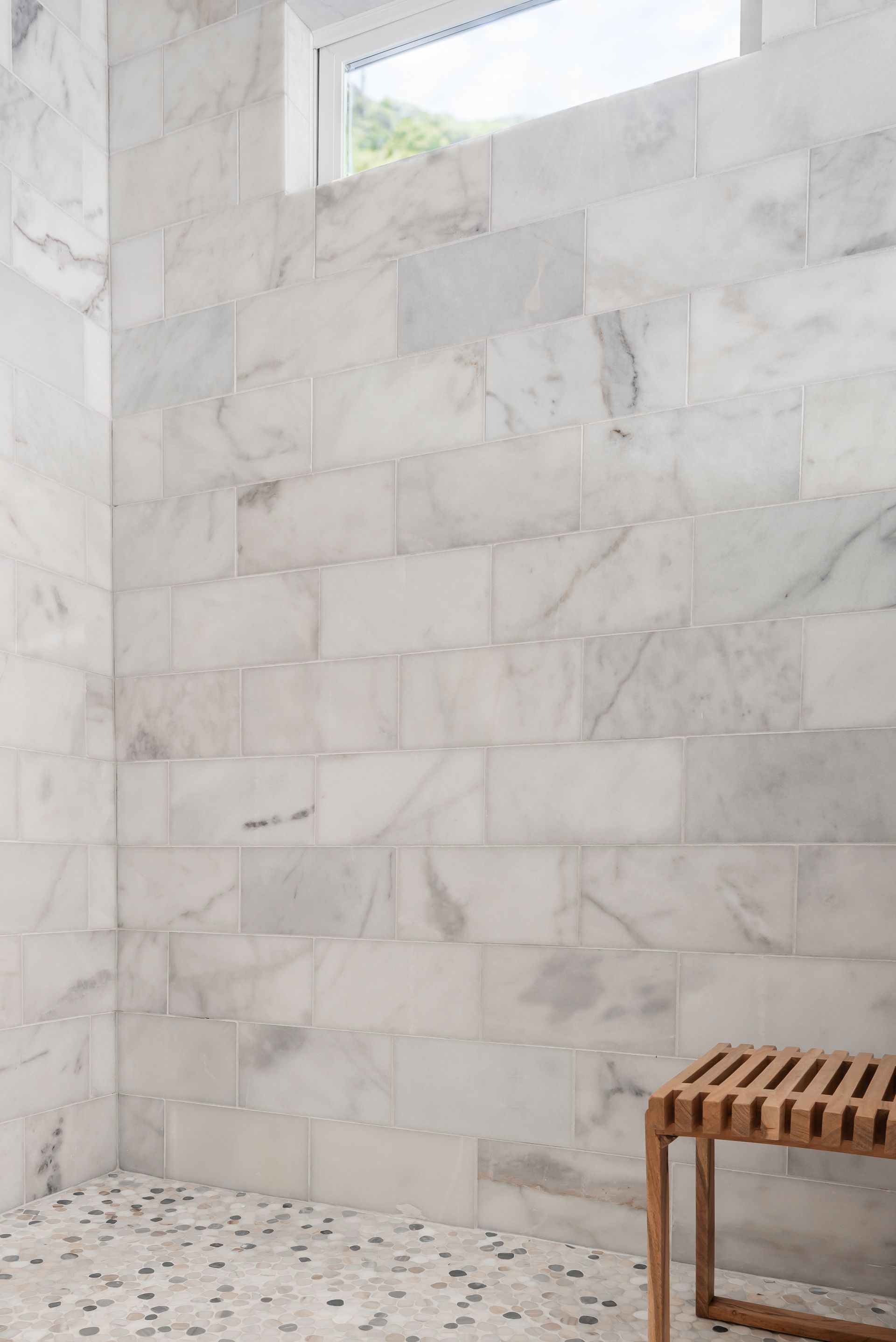 White tiled shower wall with a small frosted window and a wooden bench in the corner