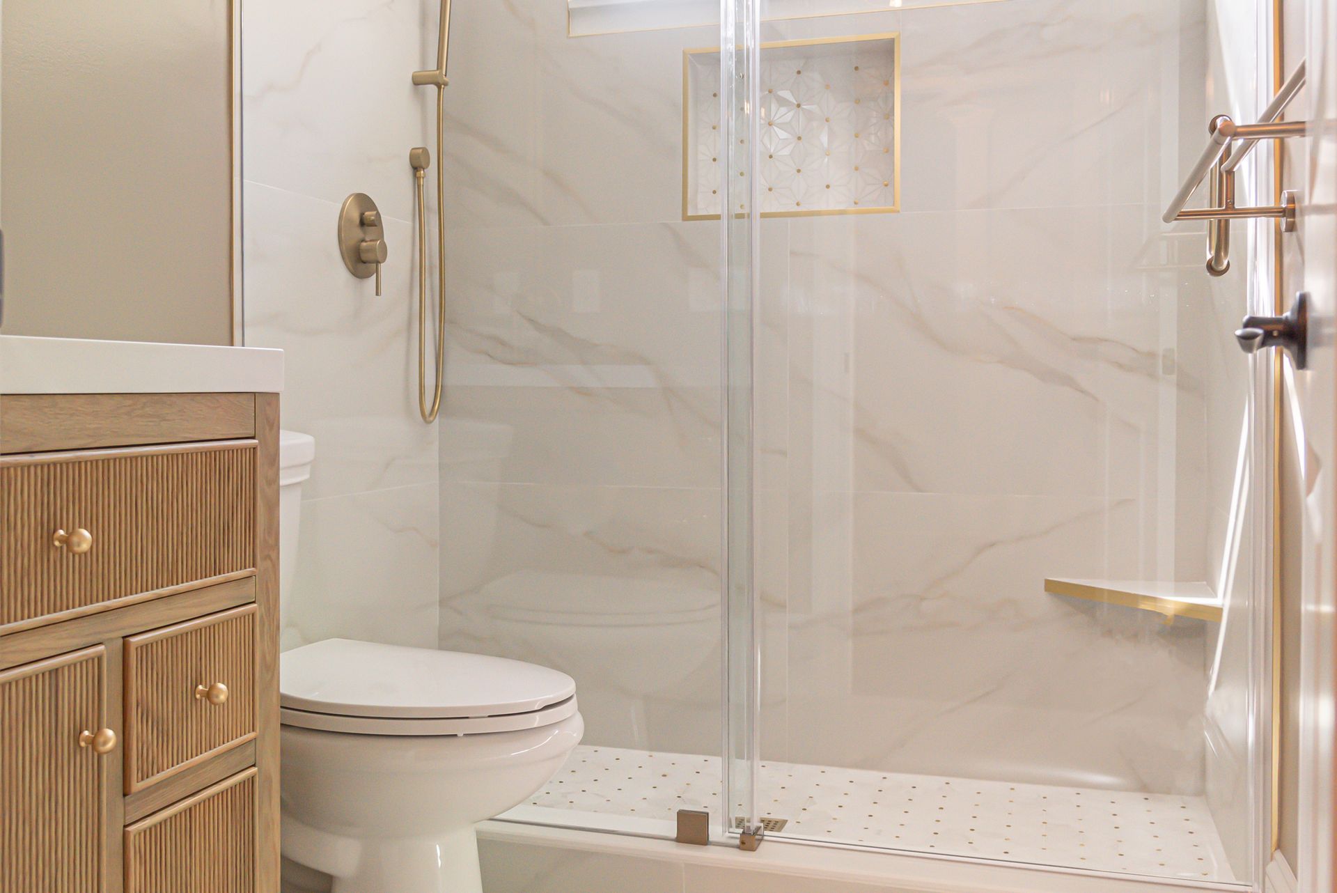 Modern bathroom with a glass shower, white tile walls, toilet, and wood vanity with gold accents