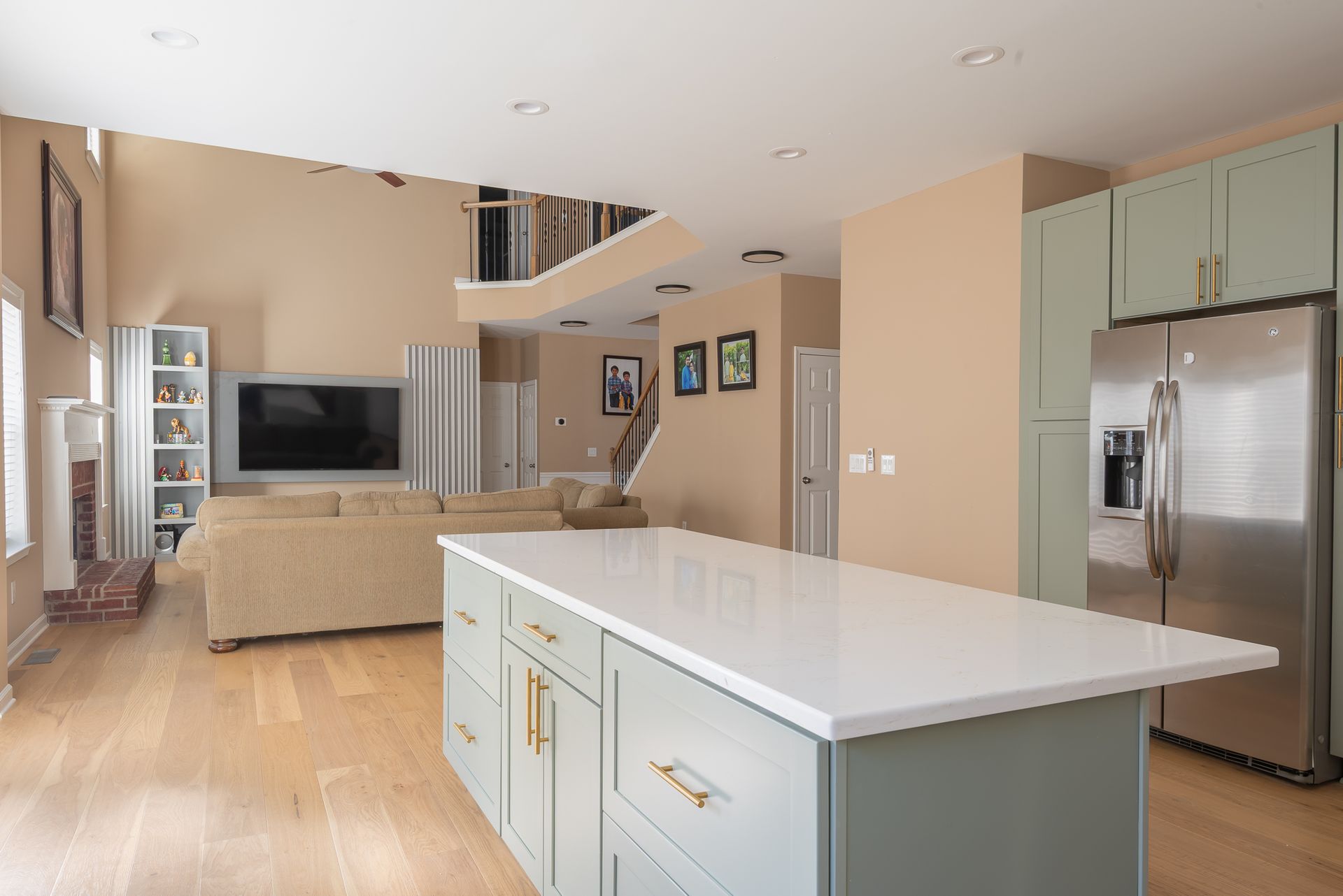 Open-plan kitchen and living room with white island, stainless fridge, beige walls, and wall-mounted TV.