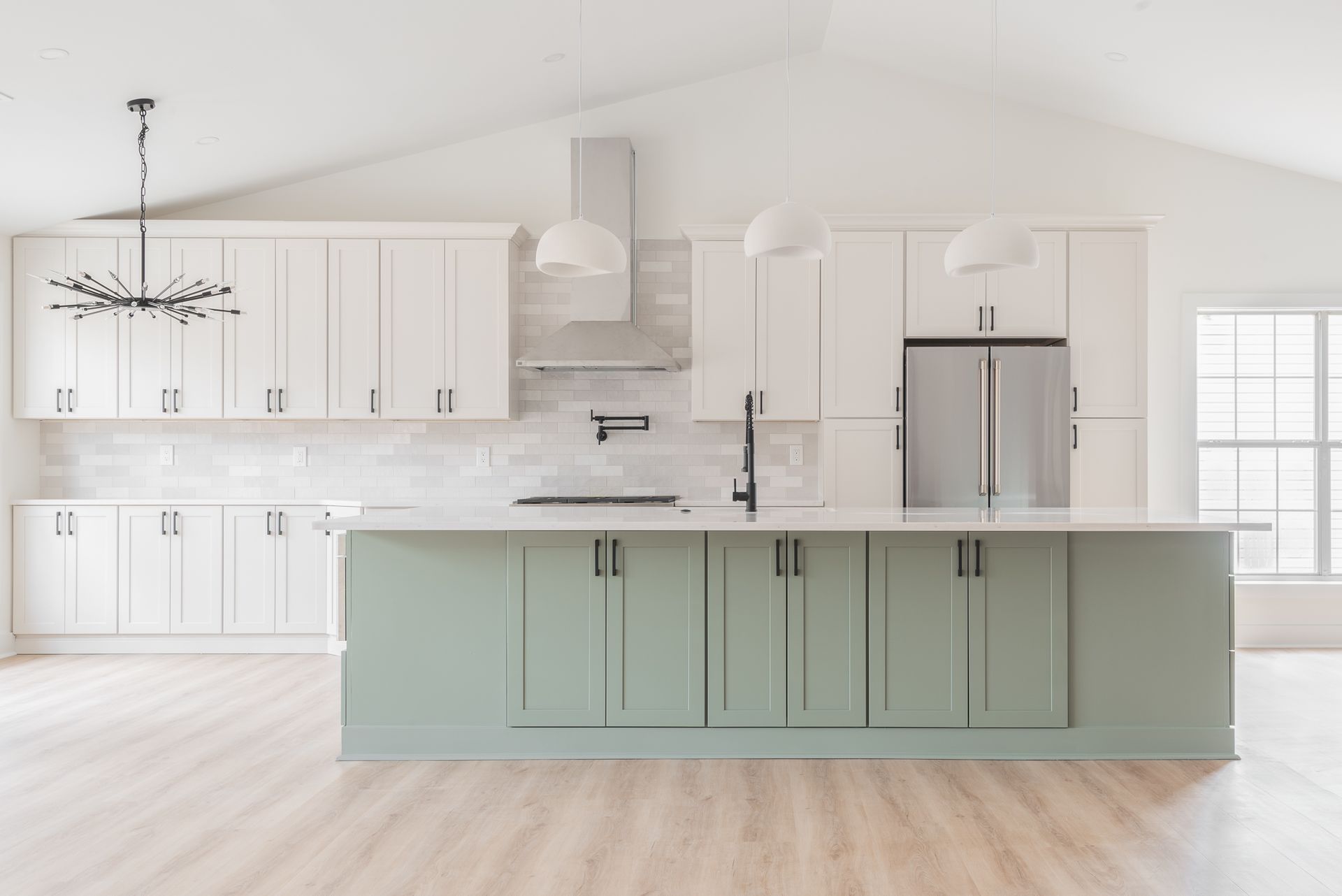 Bright white kitchen with a mint-green island, stainless fridge, and large windows