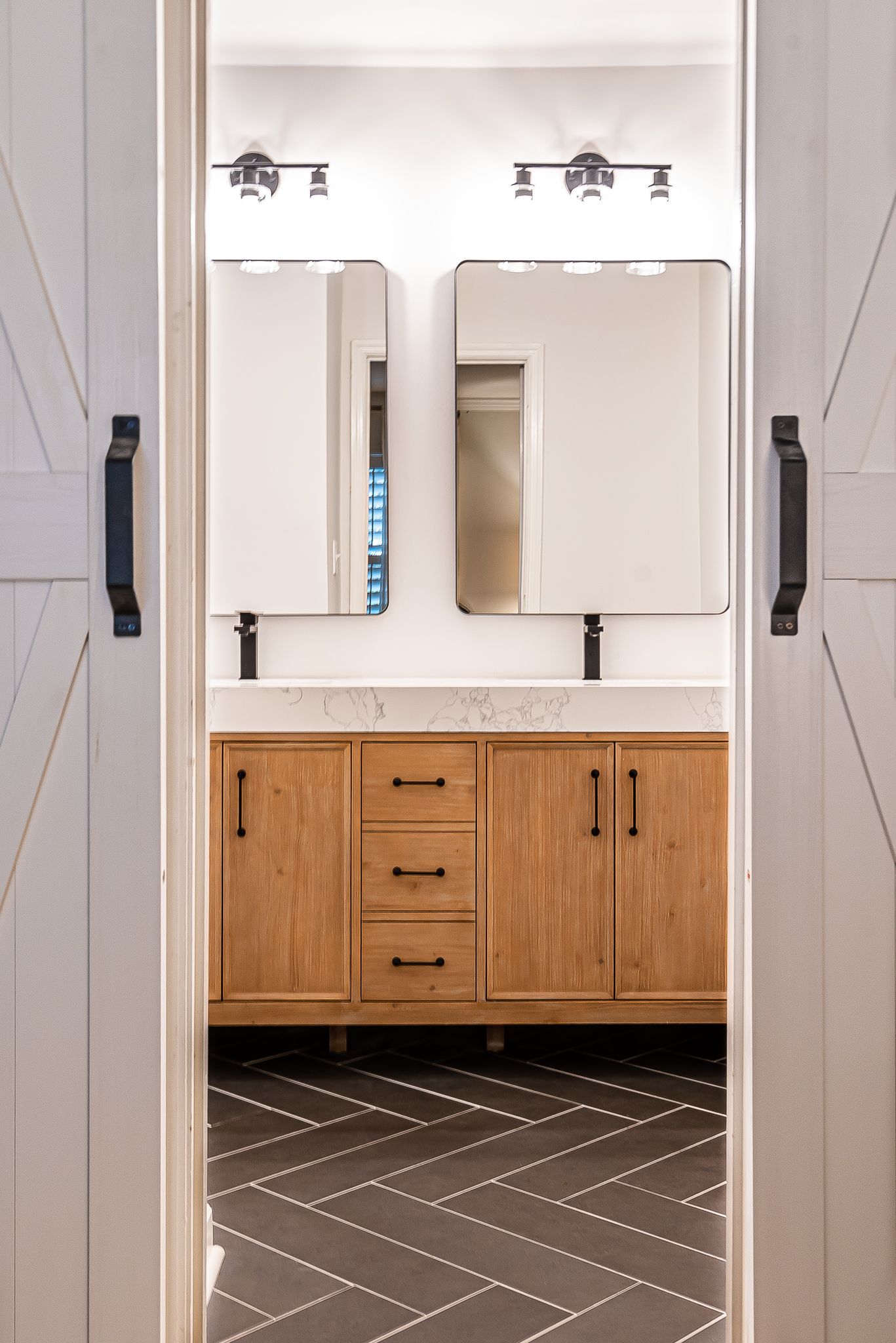 Modern bathroom vanity with double mirrors, wall sconces, and wood cabinets over patterned tile floor