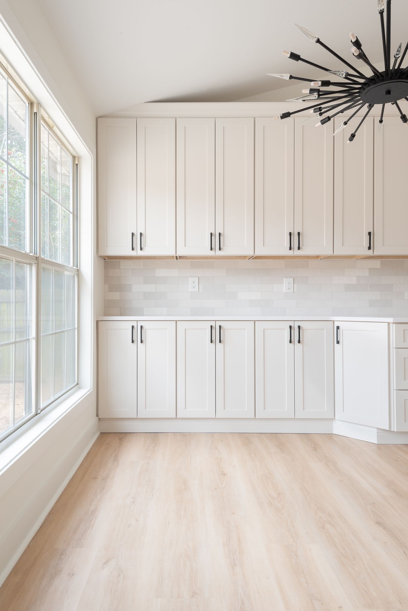 Bright laundry room with white cabinets, beige tile backsplash, light wood floor, and large window