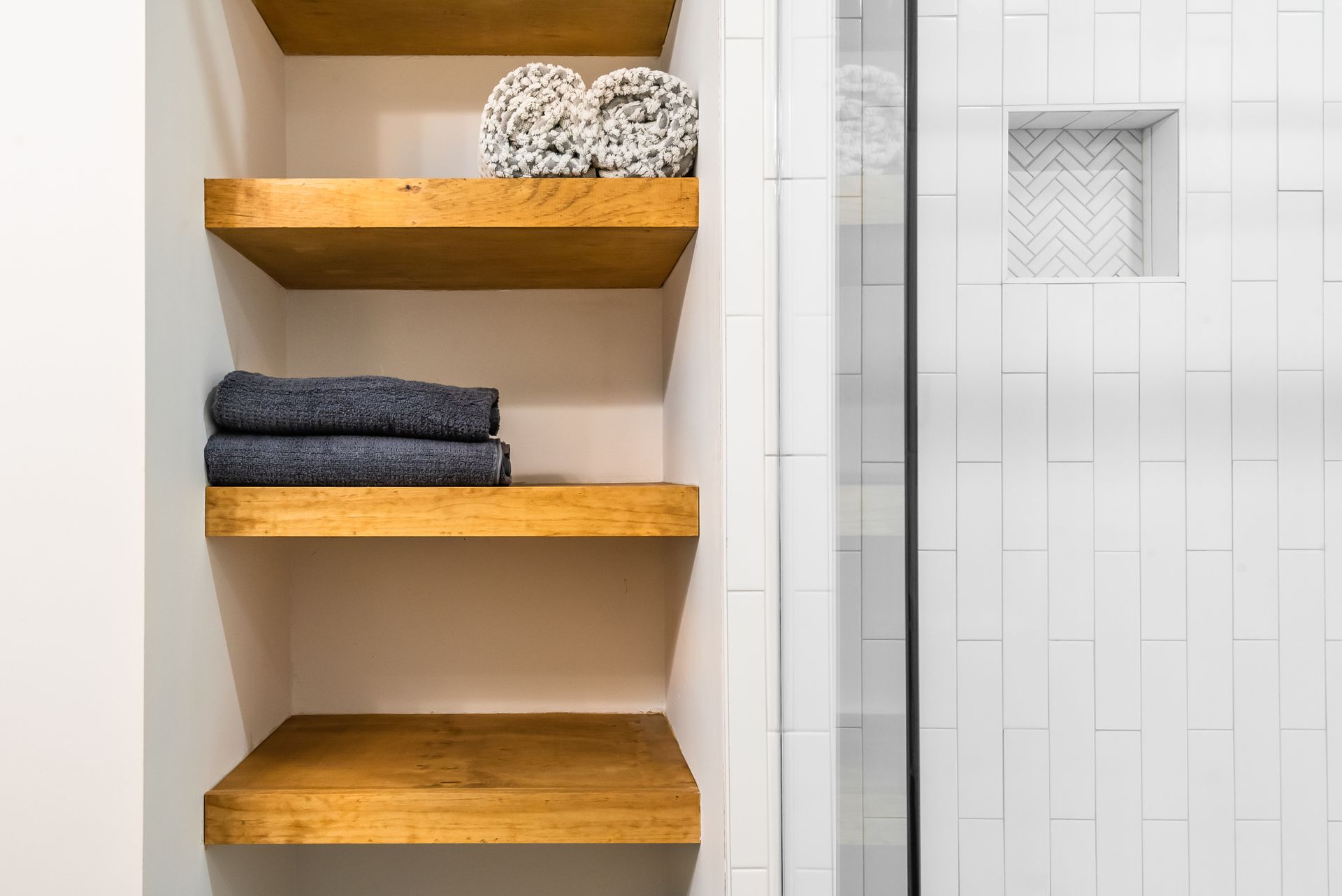 Wooden bathroom shelves with folded towels beside a tiled shower door
