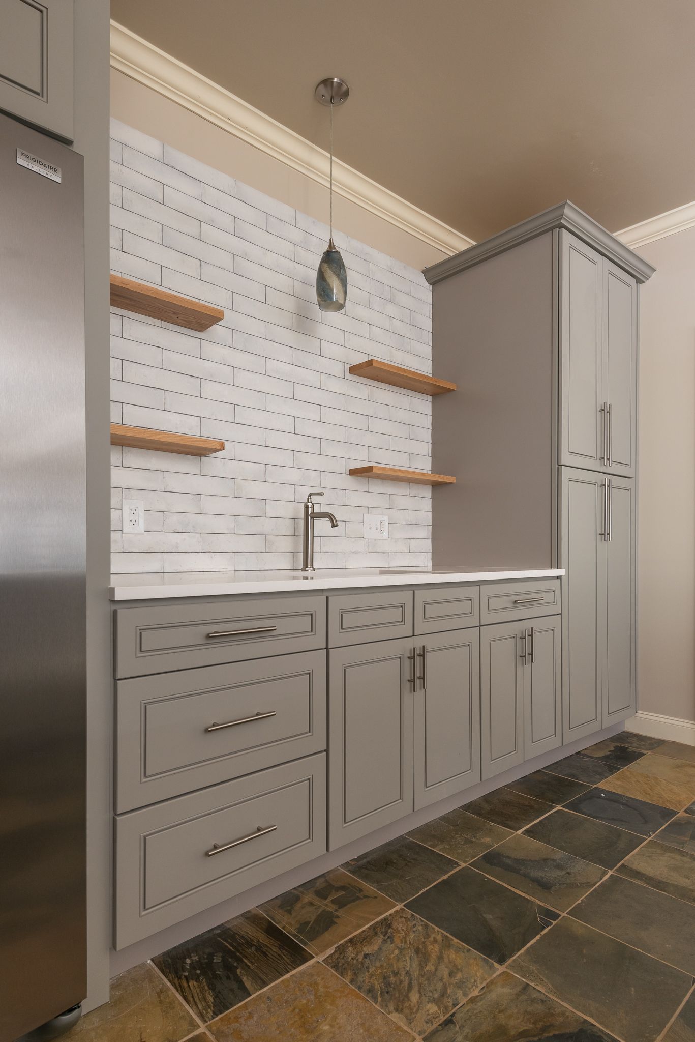 Gray kitchen with white tile backsplash, wood shelves, and tall pantry cabinets on a stone floor