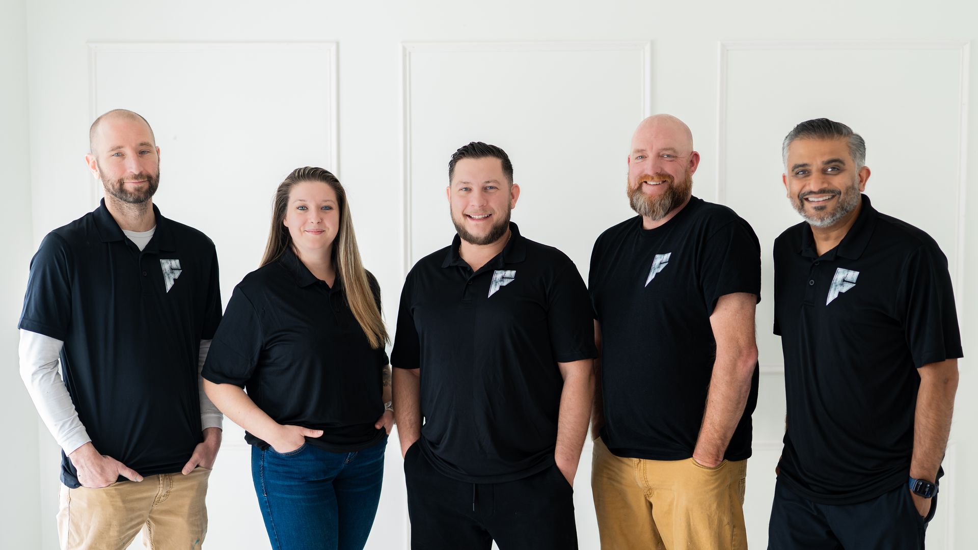 Five people in black shirts standing together indoors against a white wall.