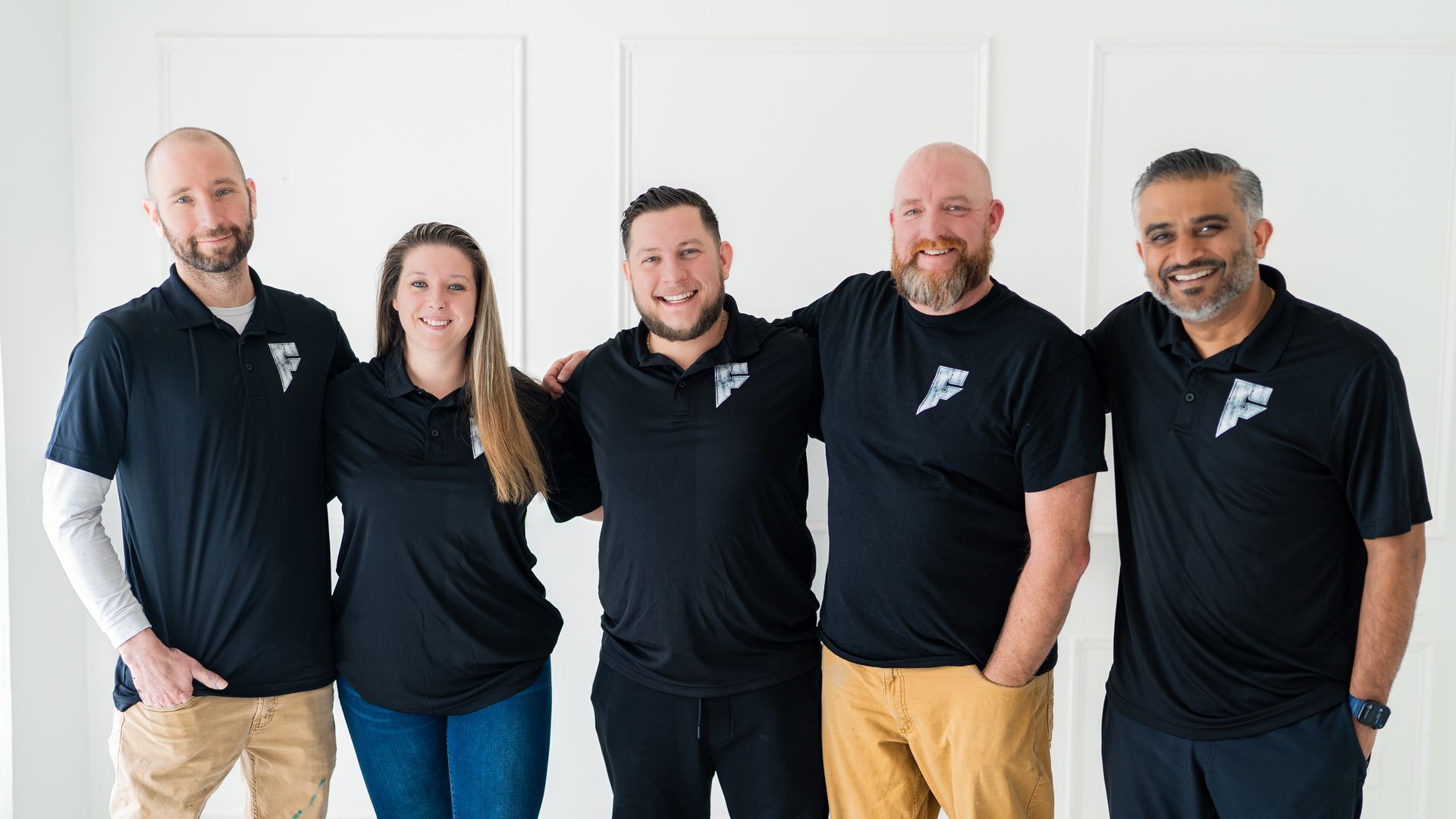 Five coworkers in black polos posing together against a white wall, smiling at the camera.