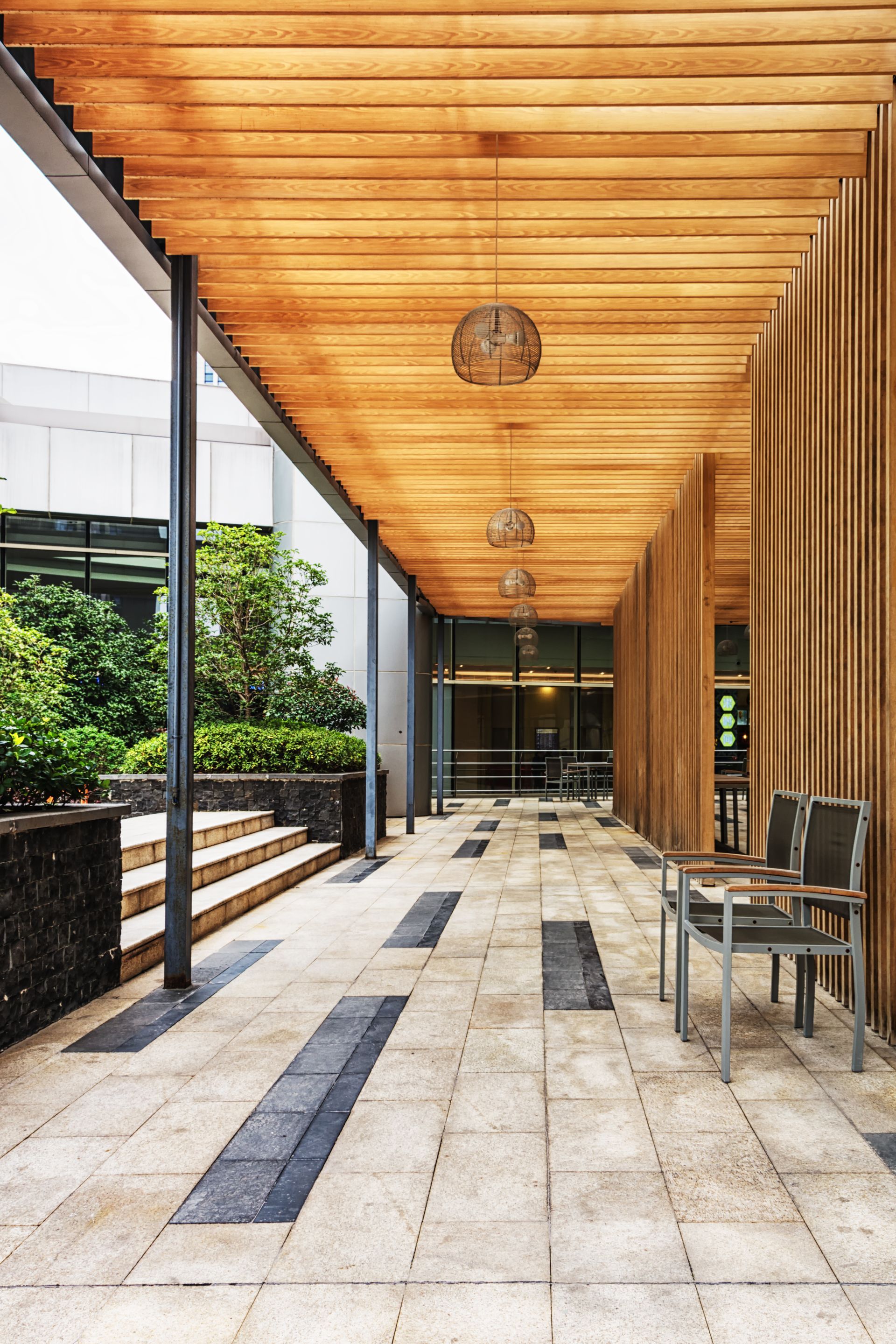 Modern covered walkway with wooden ceiling, stone floor, potted plants, and a chair by glass doors