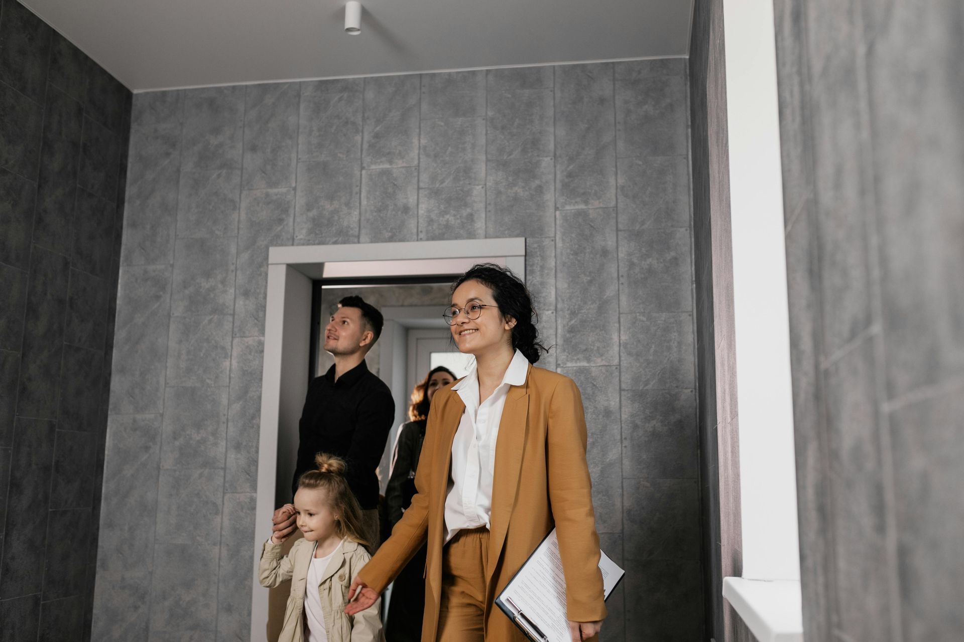 Three people stand in a gray hallway, one holding papers, with a child near the doorway.