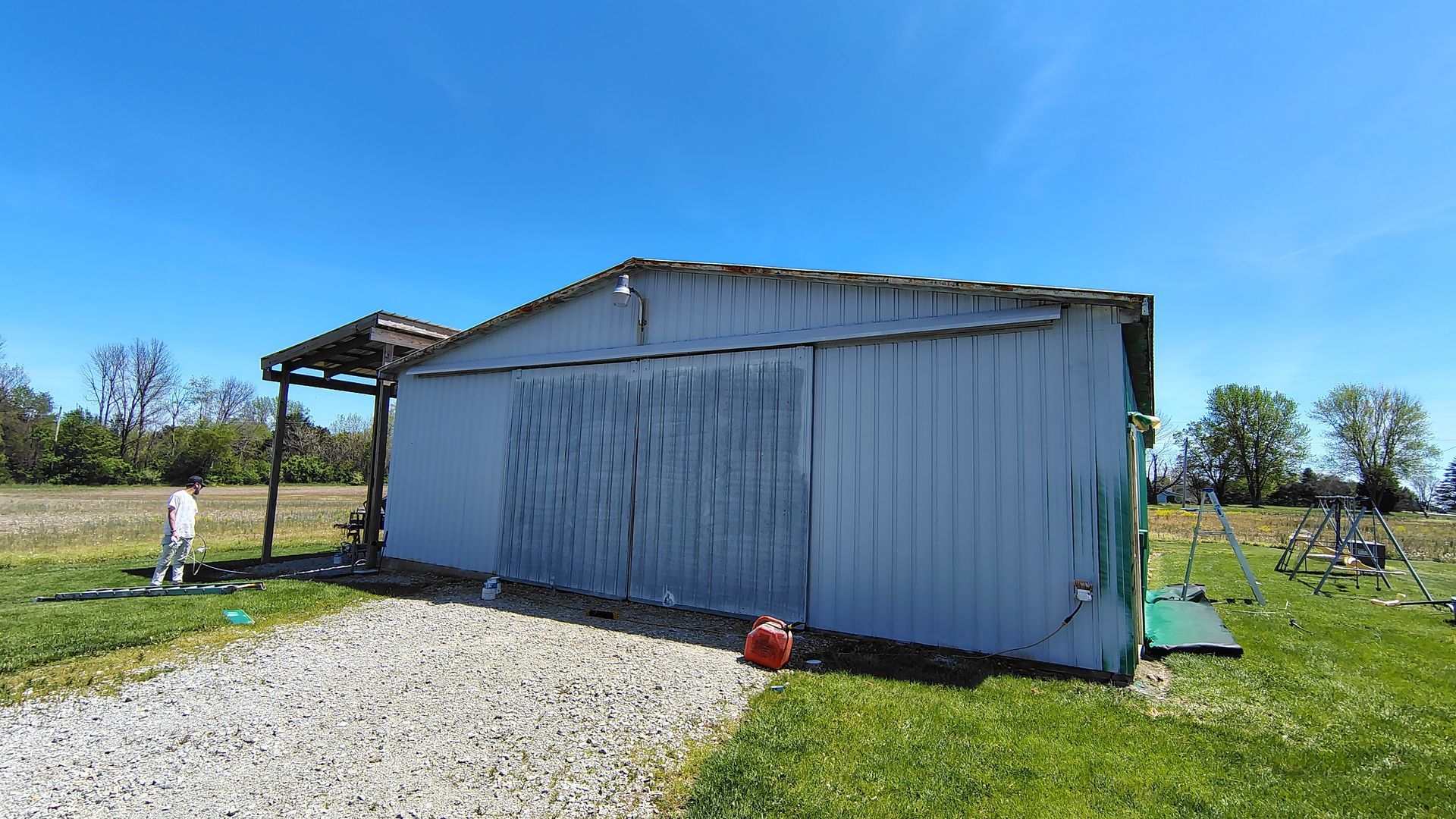 A white metal storage building with sliding doors, a small open shelter on the left, and a gravel driveway in a grassy lot.