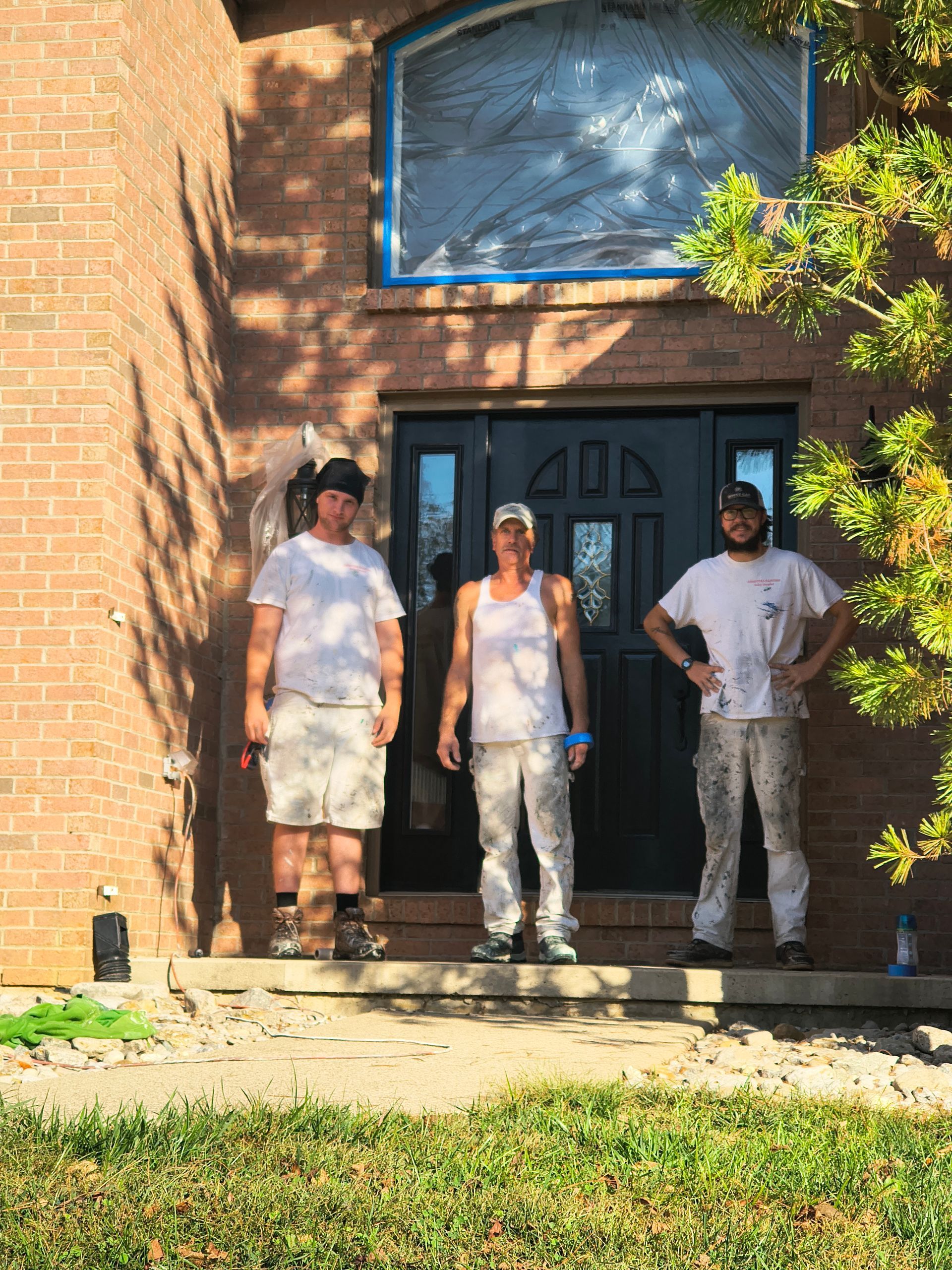Three individuals stand smiling on the front porch of a brick home with a newly painted black door.