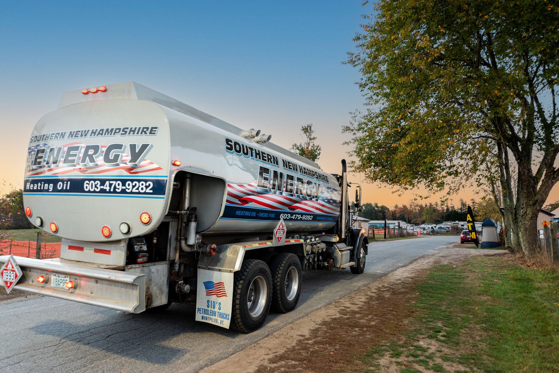 White fuel tanker truck parked on a roadside at sunset, with trees and parked cars nearby
