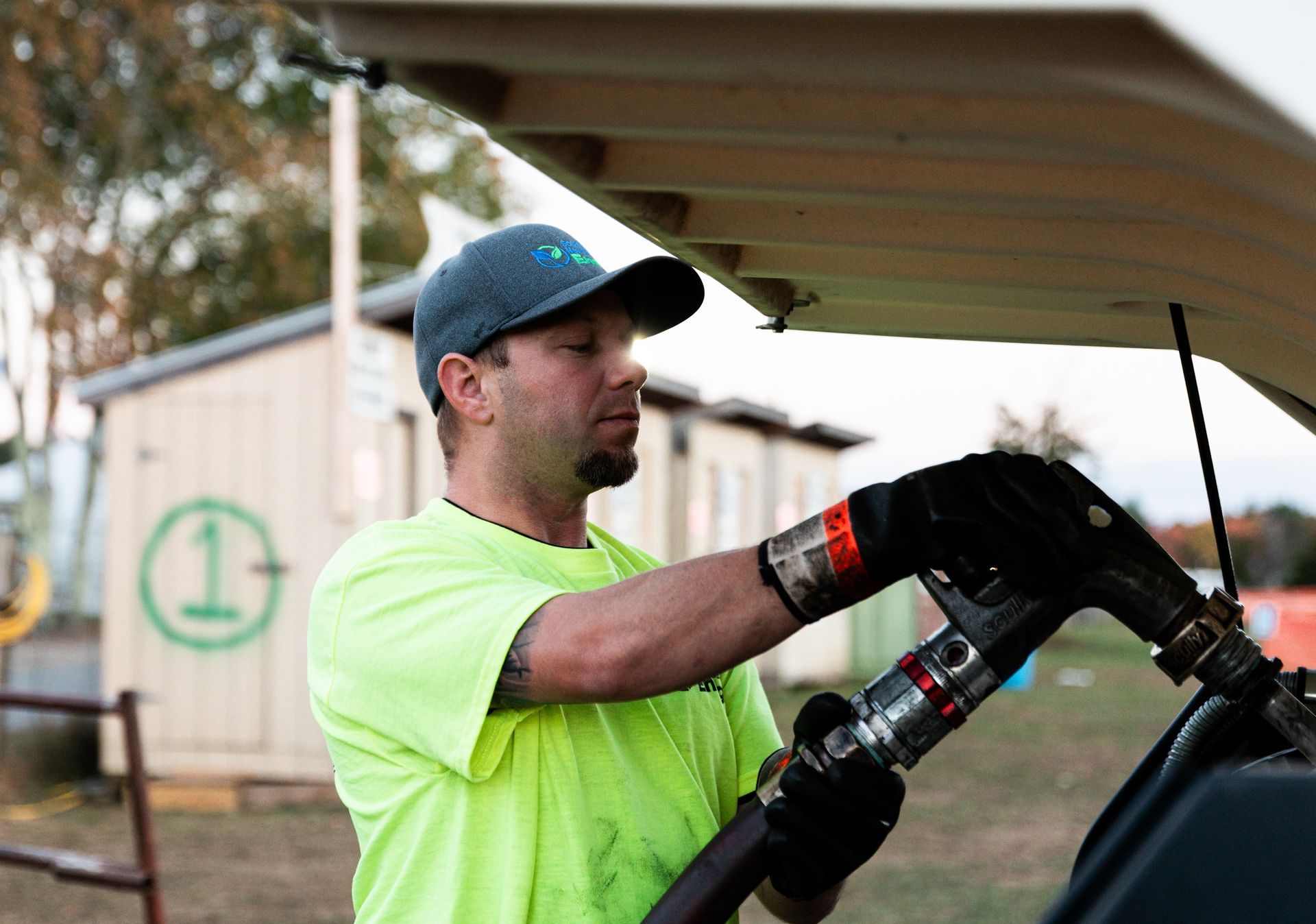 Man in neon shirt fueling a vehicle outdoors, near a small building.