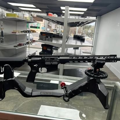 Black rifle displayed on a gun rest in a store with ammunition on shelves in the background.