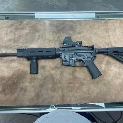 Black AR-15 rifle with red dot sight and vertical grip, lying on a brown surface.