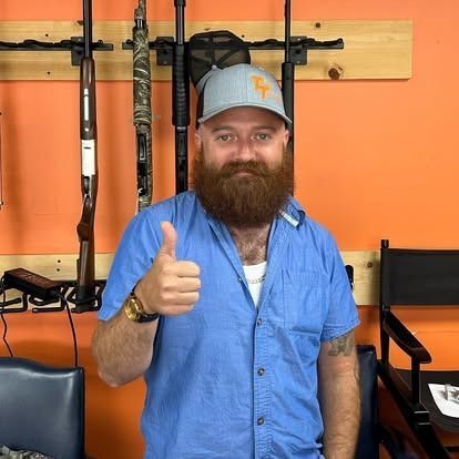 Man with beard gives thumbs up, with guns on rack in background. Orange wall.