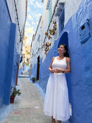 Woman in a white dress standing in a narrow blue-painted alley with hanging flowers.