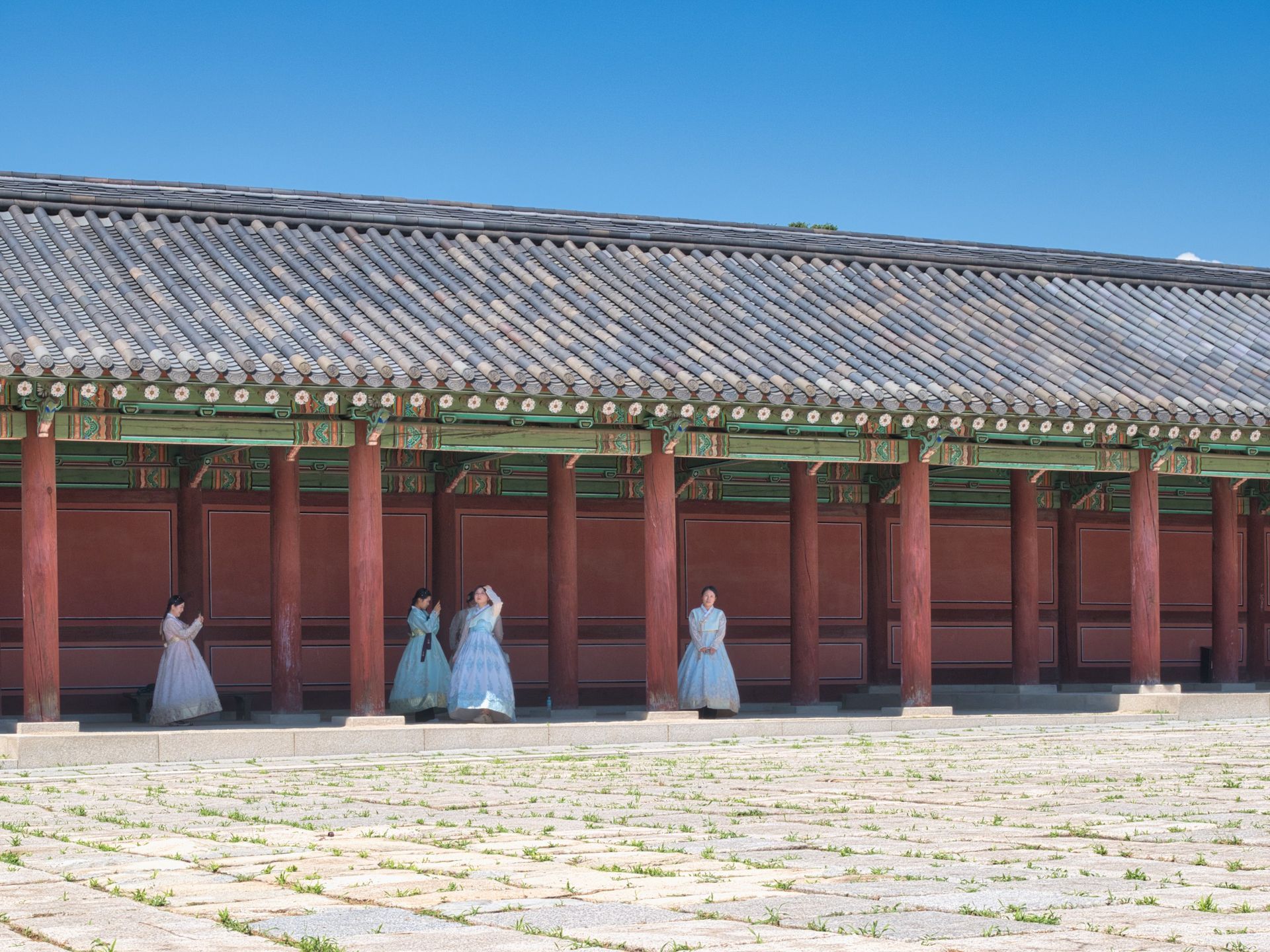 People in hanbok stand before a long red palace building with a tiled roof and blue sky.