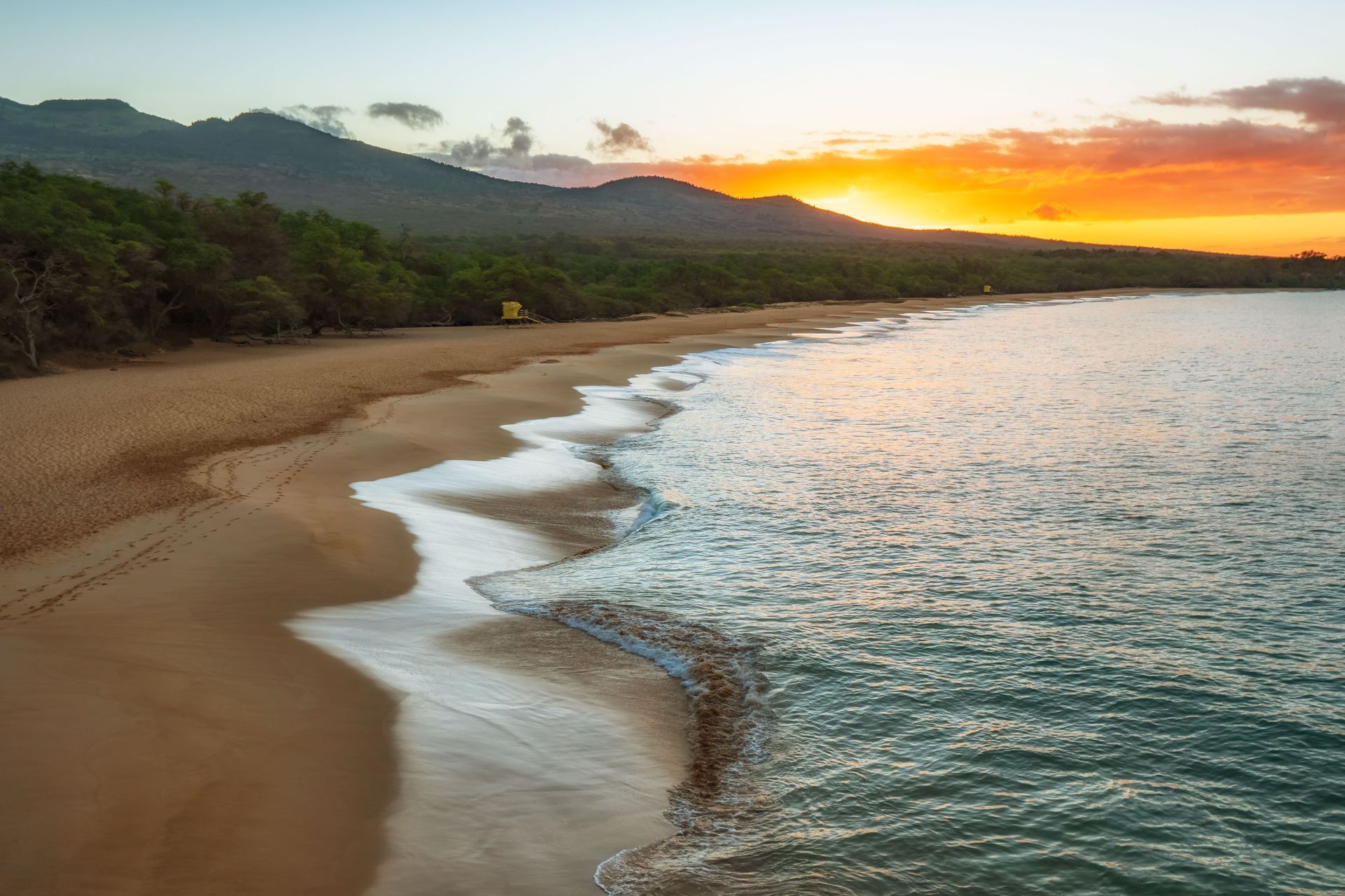 Sandy beach at sunset with gentle waves, green hills, and a glowing orange sky