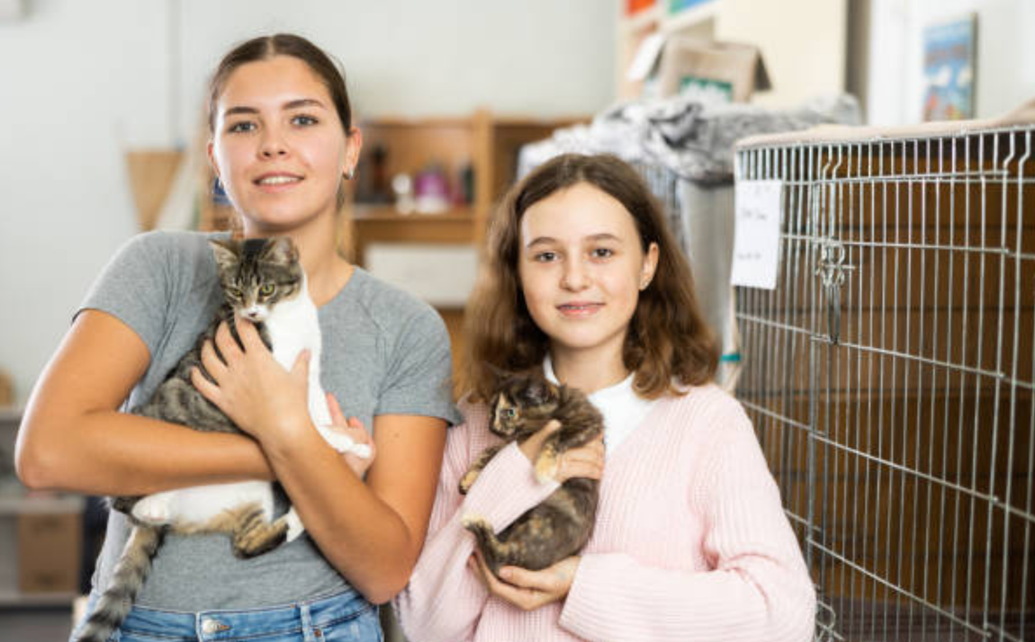 Two women holding rescued kittens beside a metal animal crate in a shelter-like room