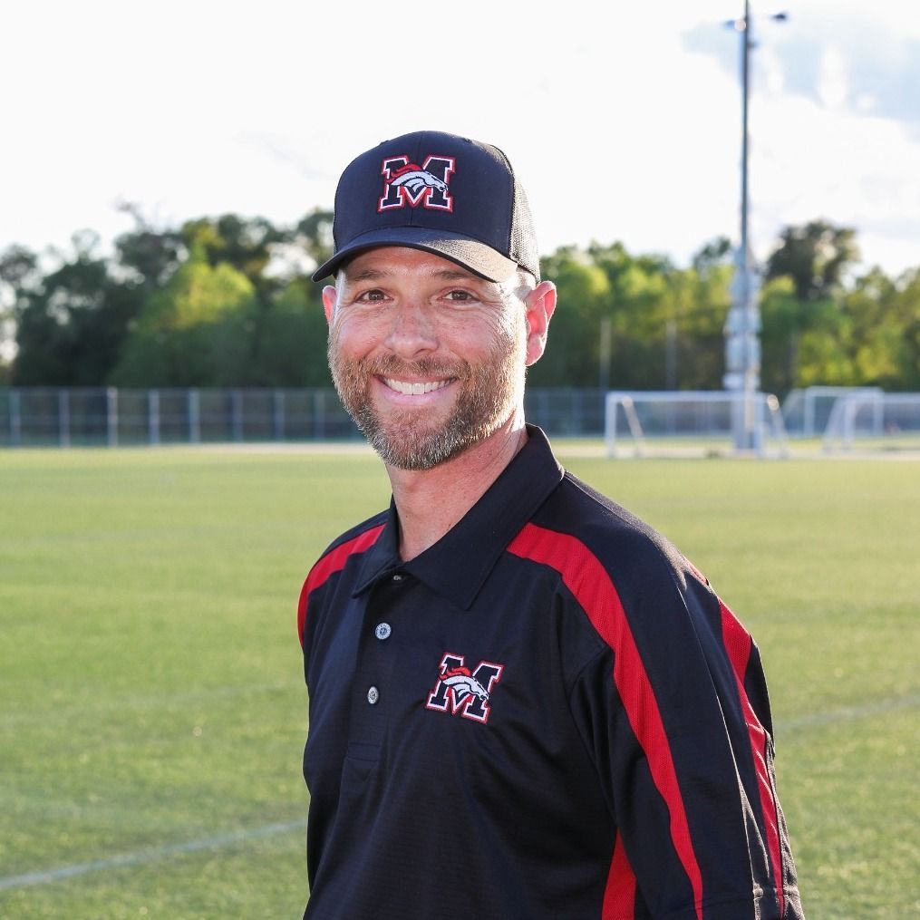 Football coach, arms crossed, on the field. Blue shirt, serious expression, players in background.