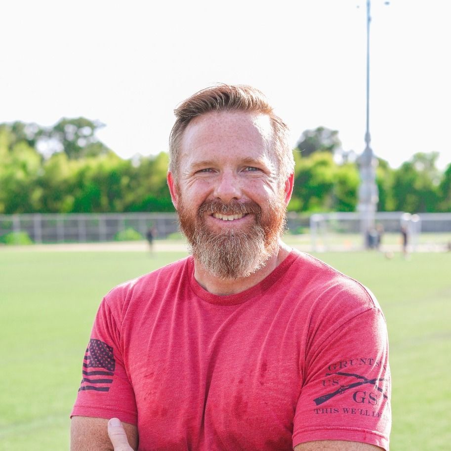 Man with crossed arms on a football field. He is wearing a blue polo shirt, khaki pants, and has a serious expression.