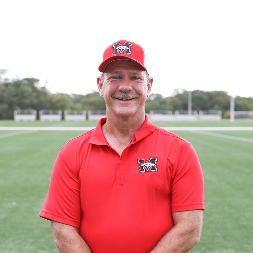 Man with crossed arms on a football field. He is wearing a blue polo shirt, khaki pants, and has a serious expression.