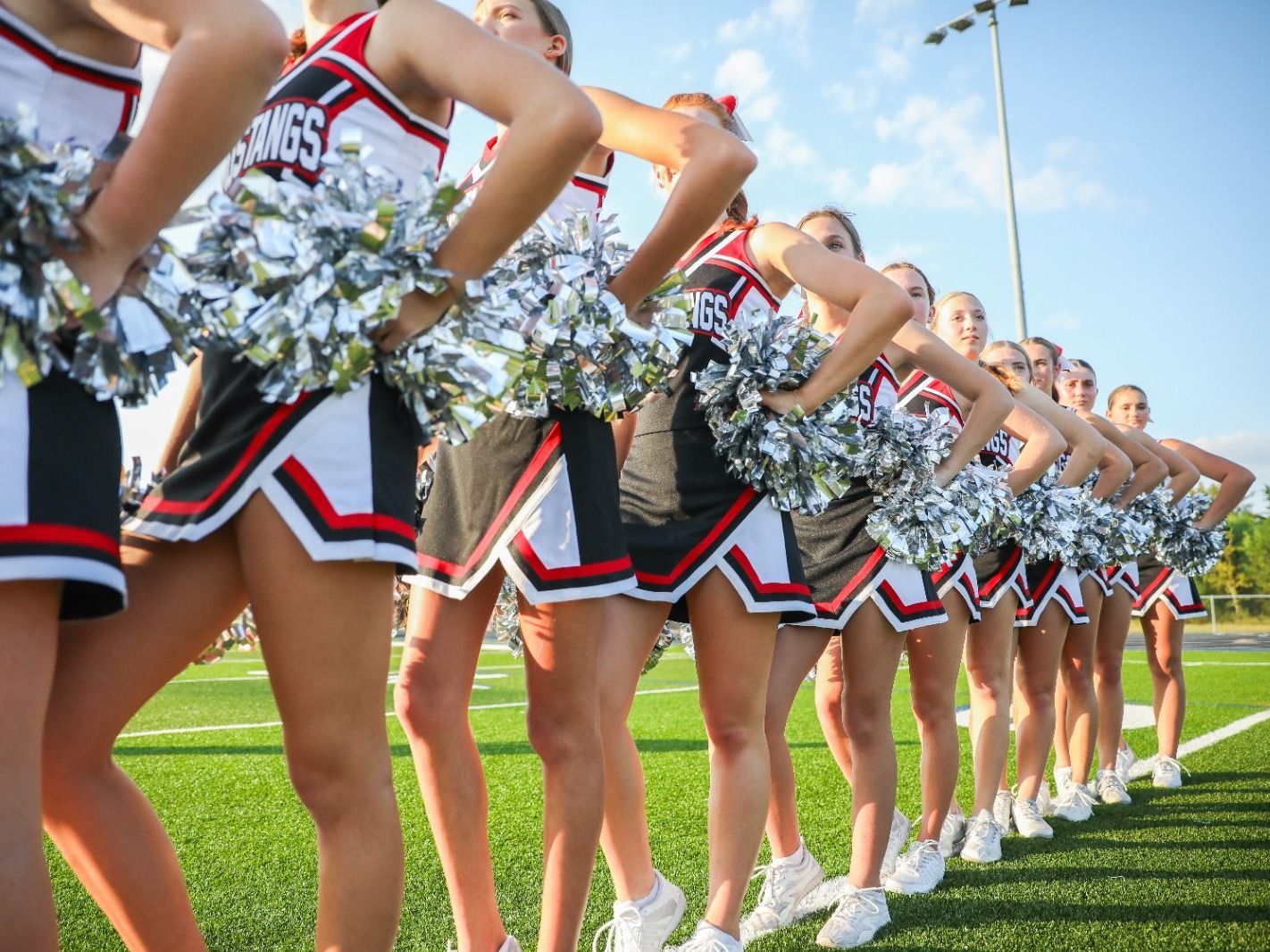 Cheerleaders in silver and red uniforms line up on a sunny football field with pom-poms raised.