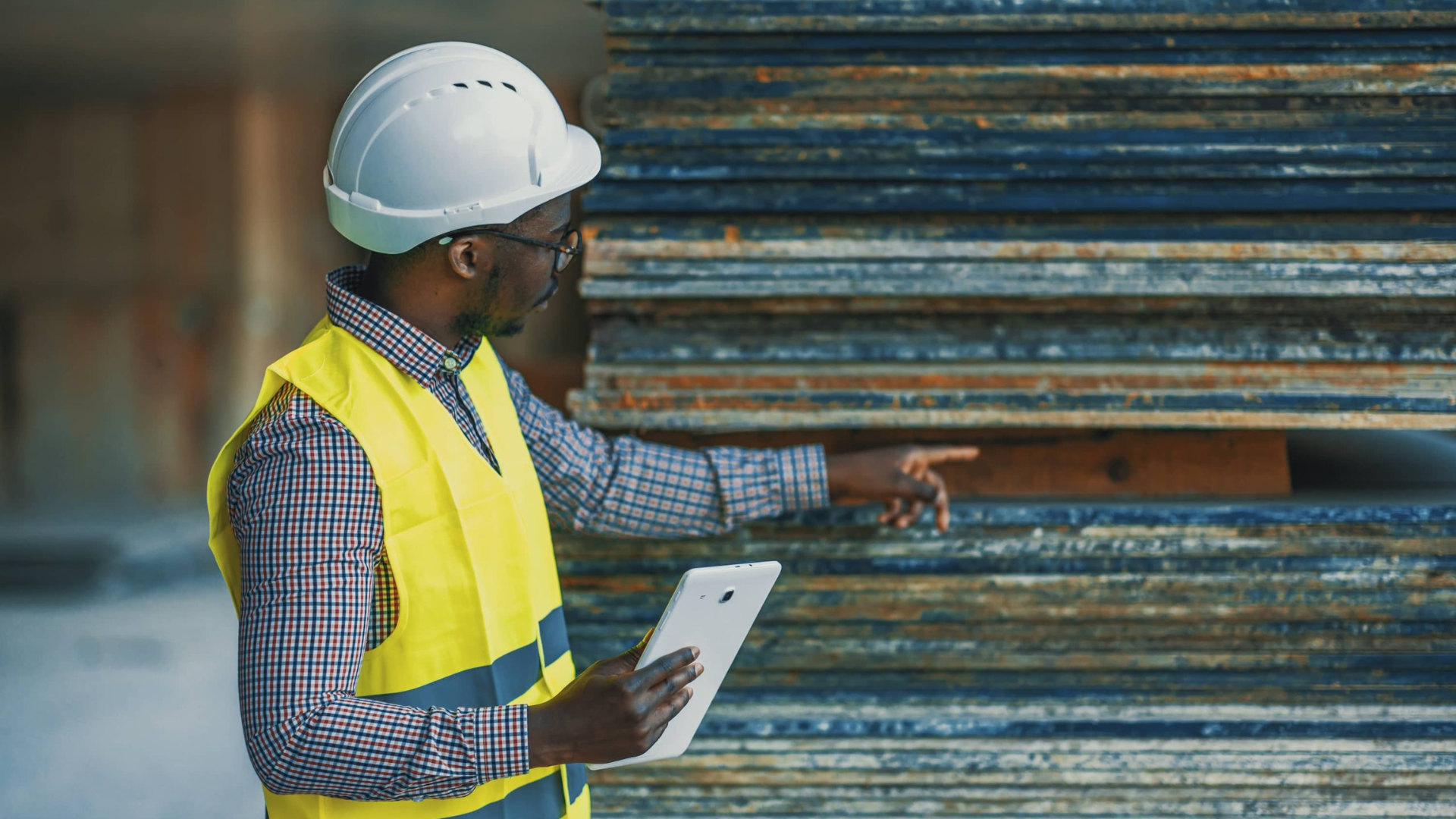 Worker in hard hat and safety vest inspecting stacked metal pipes with a clipboard.