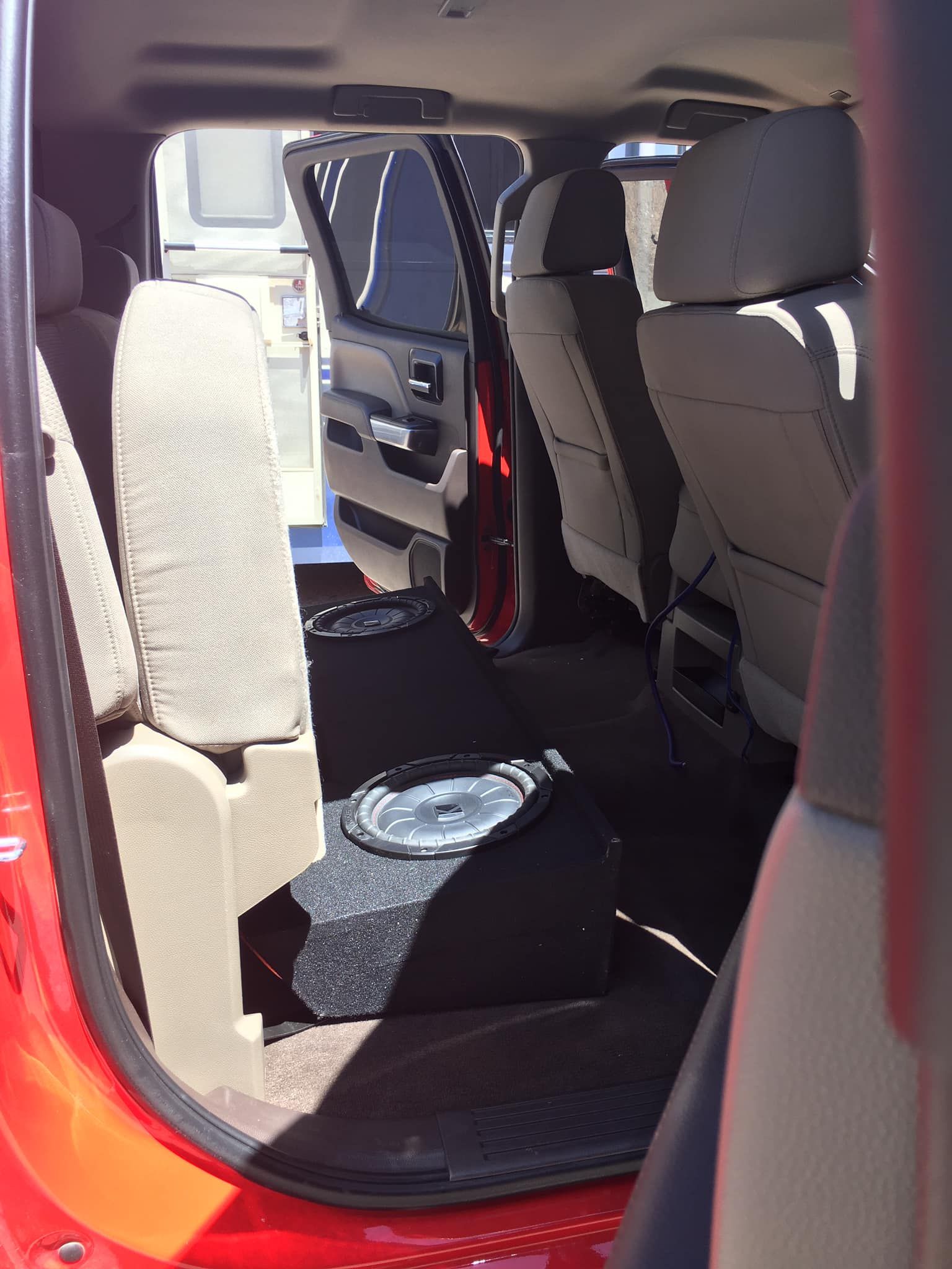 Red truck's back seat with a subwoofer enclosure. Beige seats and black carpet. Bright sunlight.