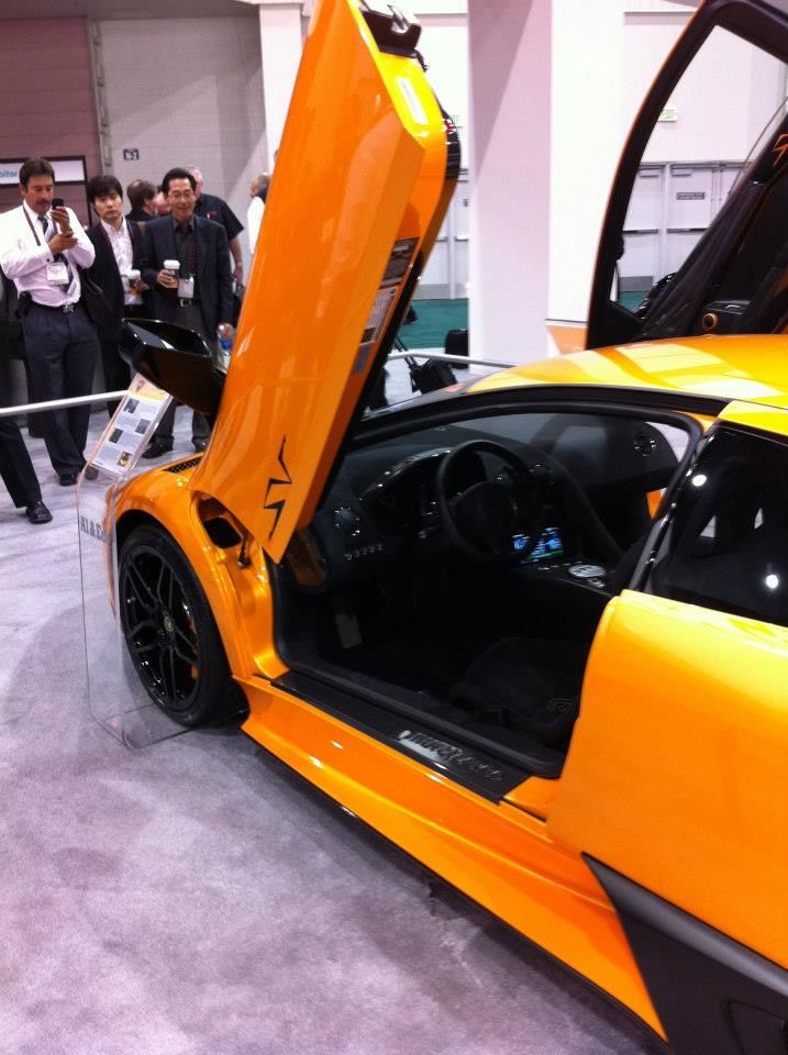 Orange Lamborghini with doors open, showing interior. People in background, indoors.
