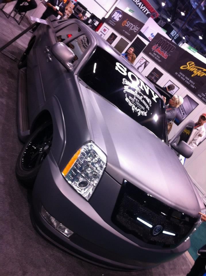 Gray Cadillac Escalade with black wheels and a Sony windshield banner at an auto show.