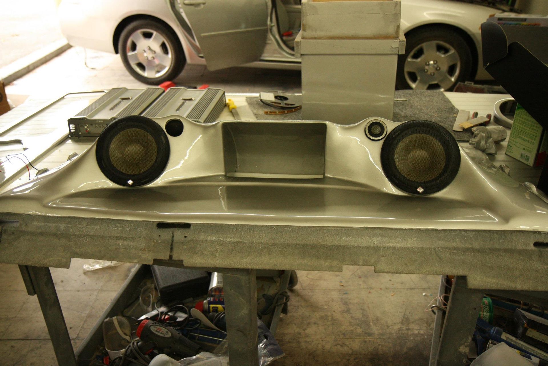 Car audio component with two speakers installed, laid out on a workbench, car visible in the background.