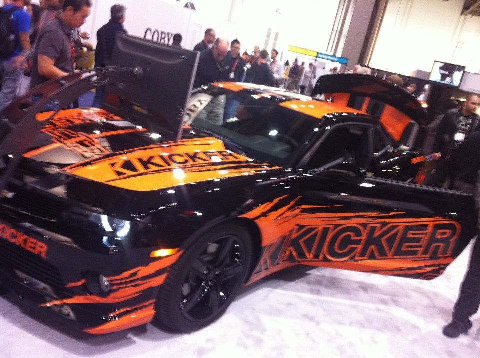 Black and orange Kicker-branded Chevrolet Camaro with open doors at an event.