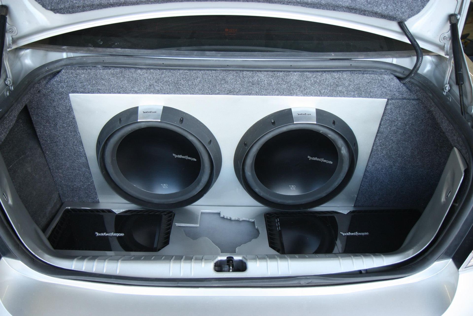 Car trunk with two large subwoofers and two amplifiers mounted on a silver panel, carpeted interior.