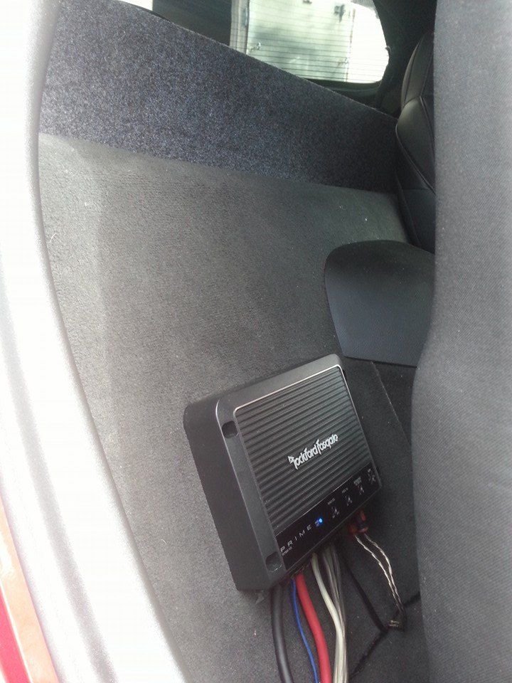 A car audio amplifier, likely Rockford Fosgate, mounted behind a car seat with various wires.
