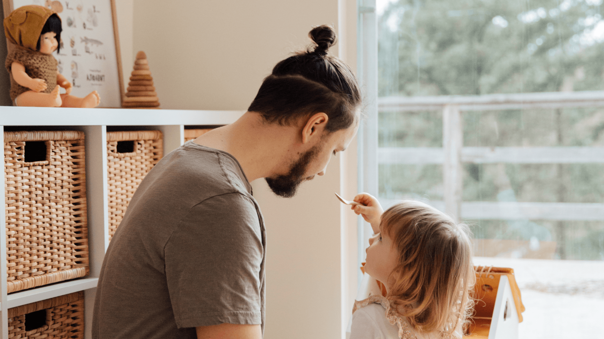 Adult and child face each other indoors, sharing a playful moment near a bright window.