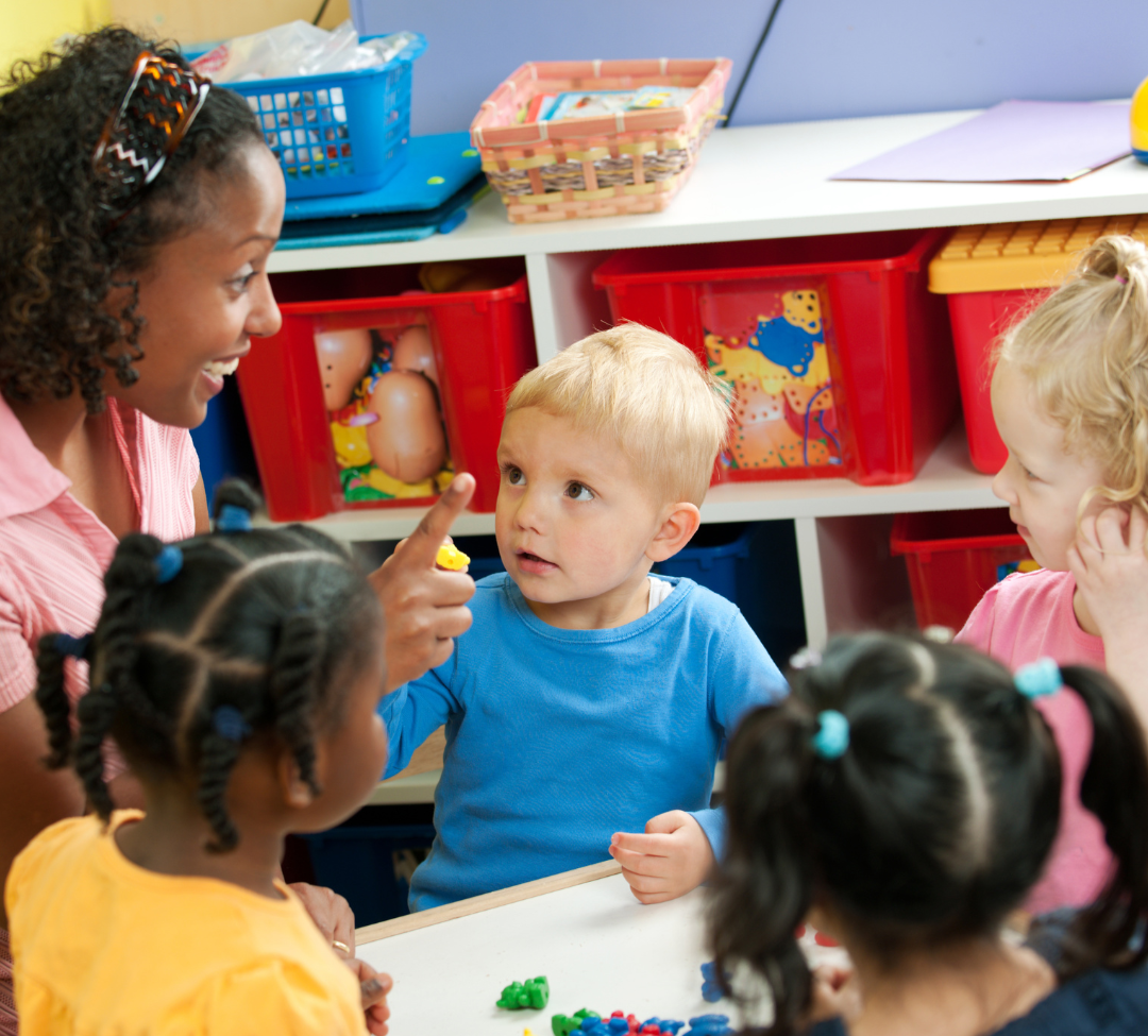 Teacher and young children gathered around a table in a colorful classroom, talking and playing with toys