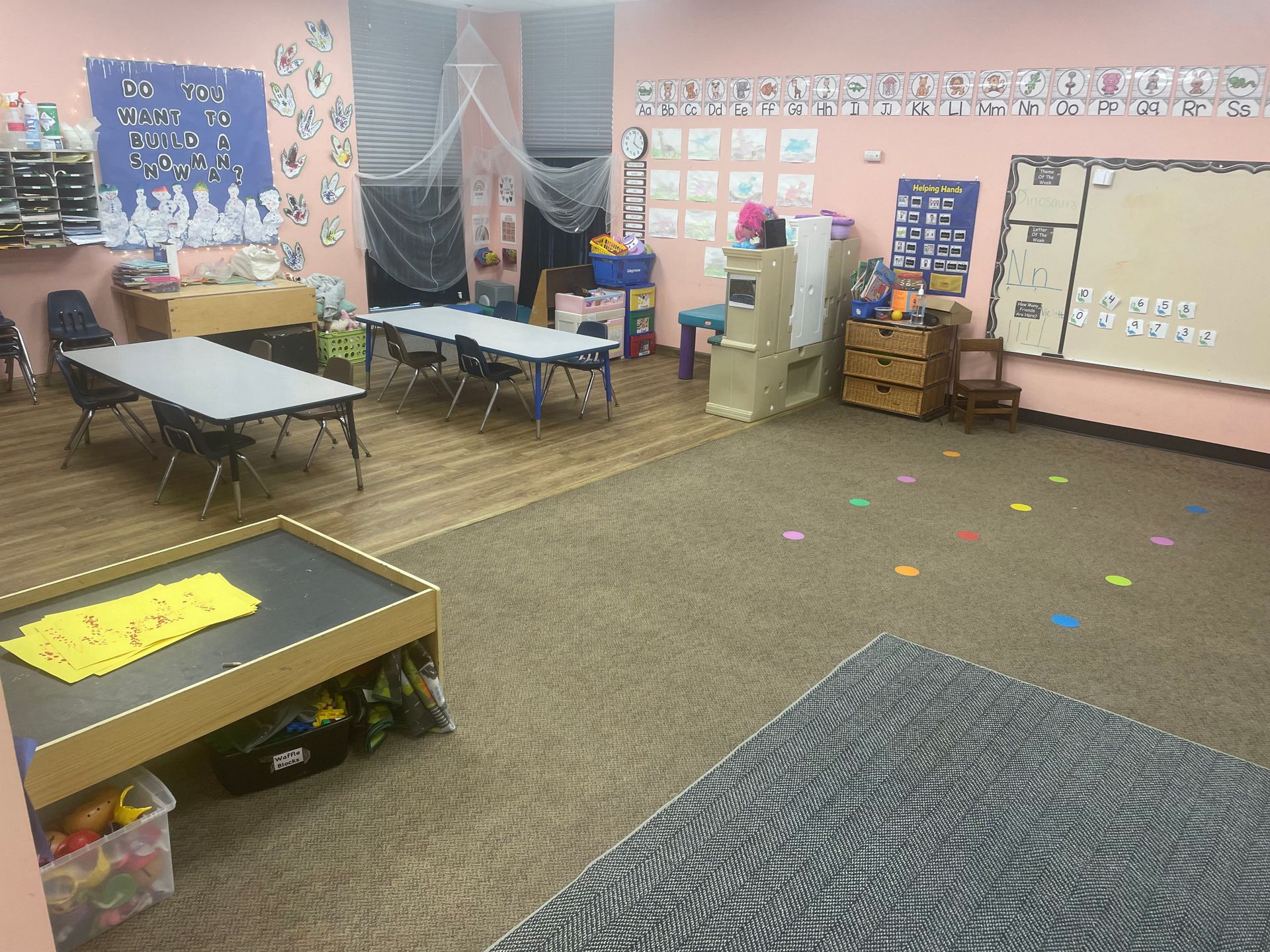 Empty classroom with small tables, carpeted floor, bulletin boards, and colorful posters on the walls.