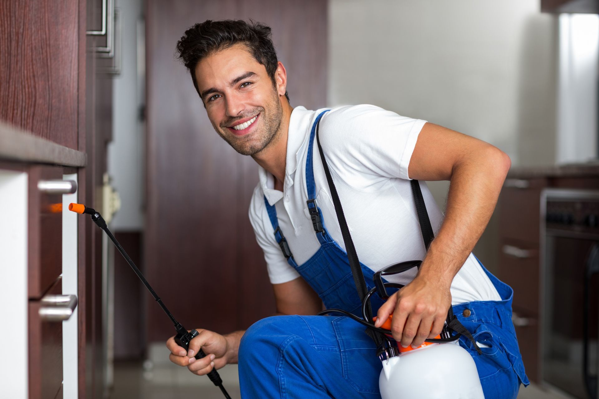Worker in blue overalls spraying a red wall with a paint sprayer, smiling in a hallway.