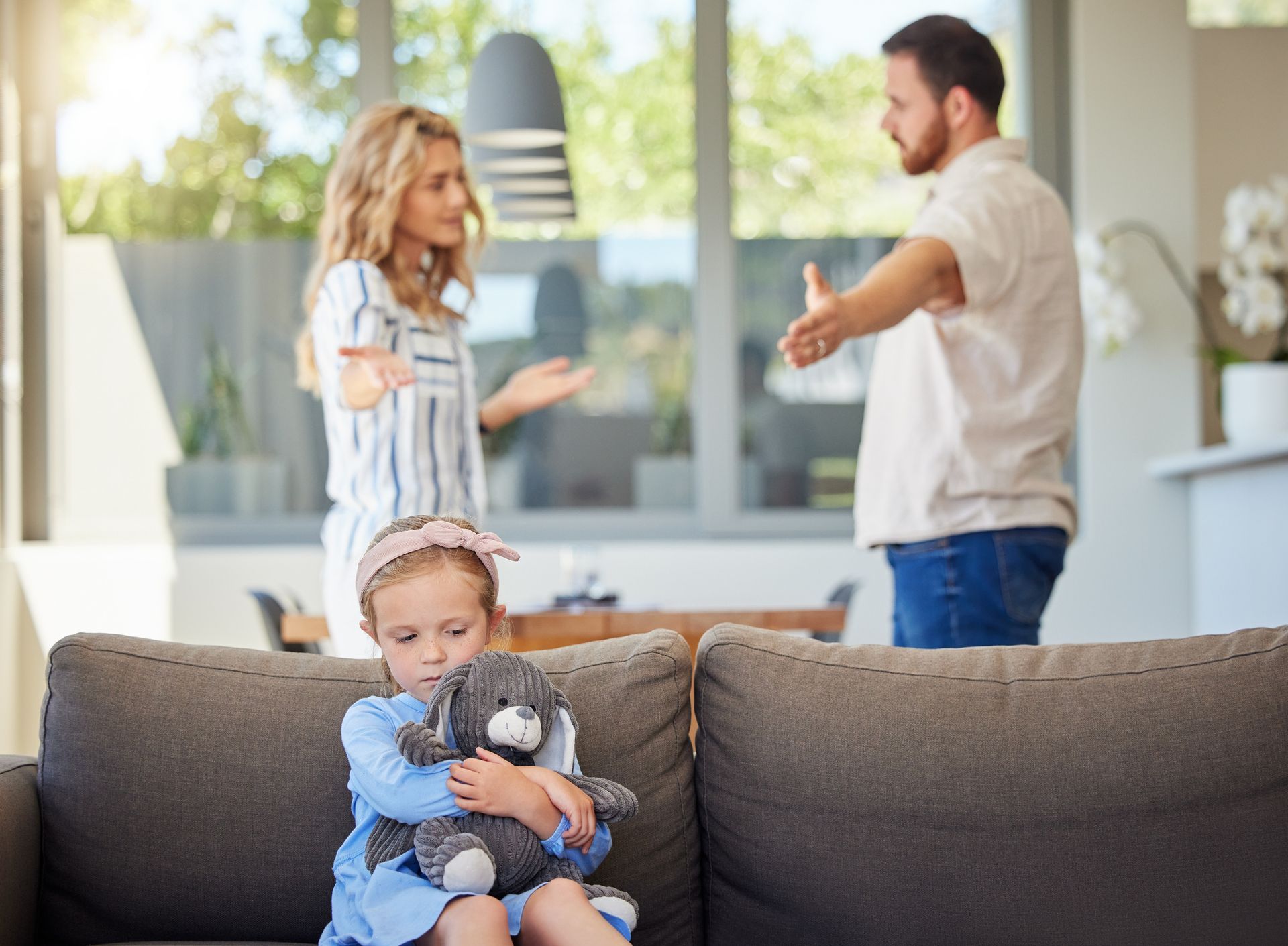 Girl hugging teddy bear, parents arguing in background, living room.