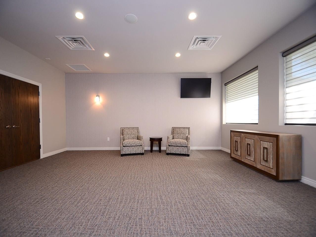Empty carpeted room with two chairs, wall-mounted TV, windows, and a side cabinet