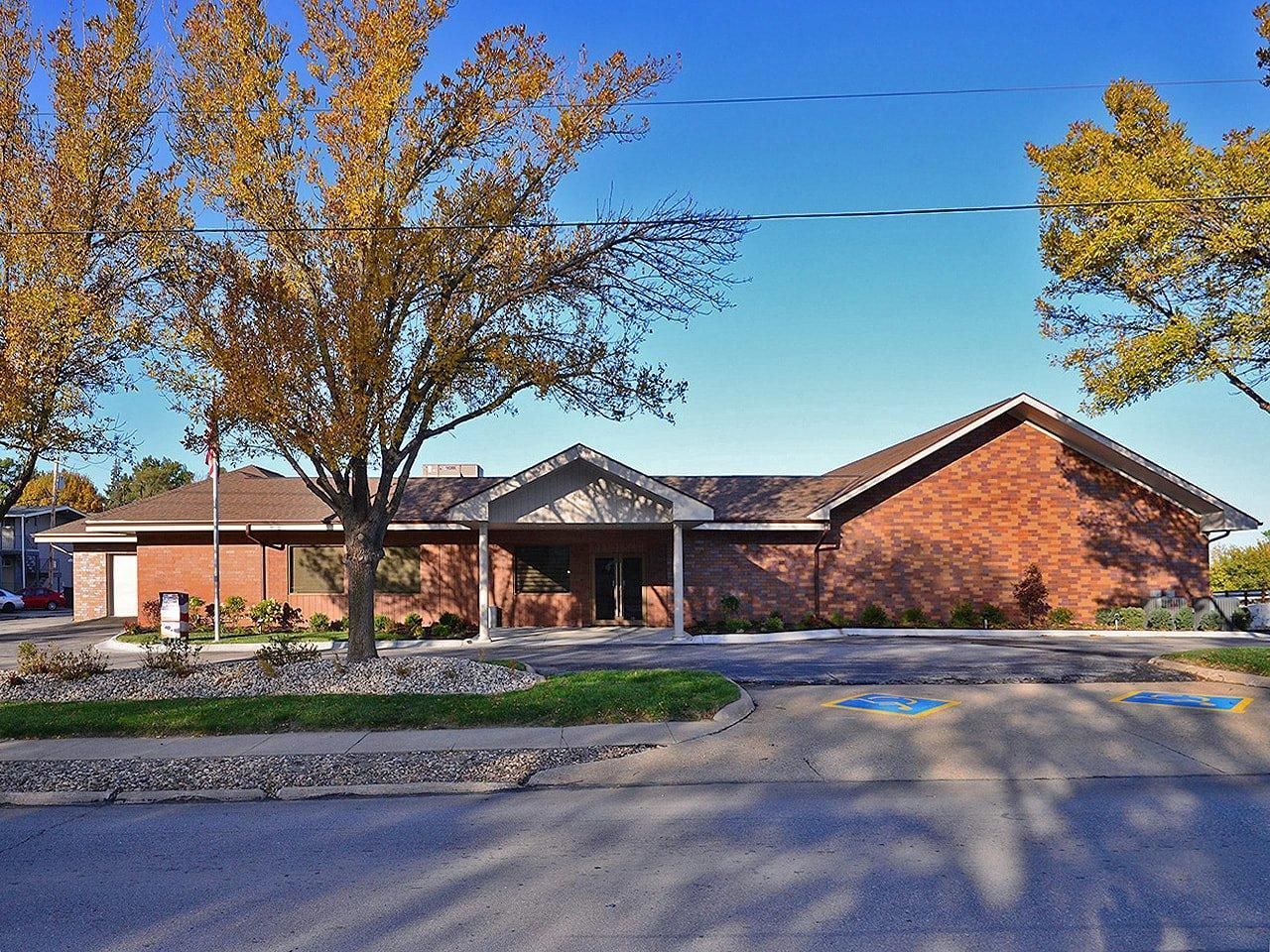Brick building with a gabled roof and autumn trees in front, seen from a parking lot.